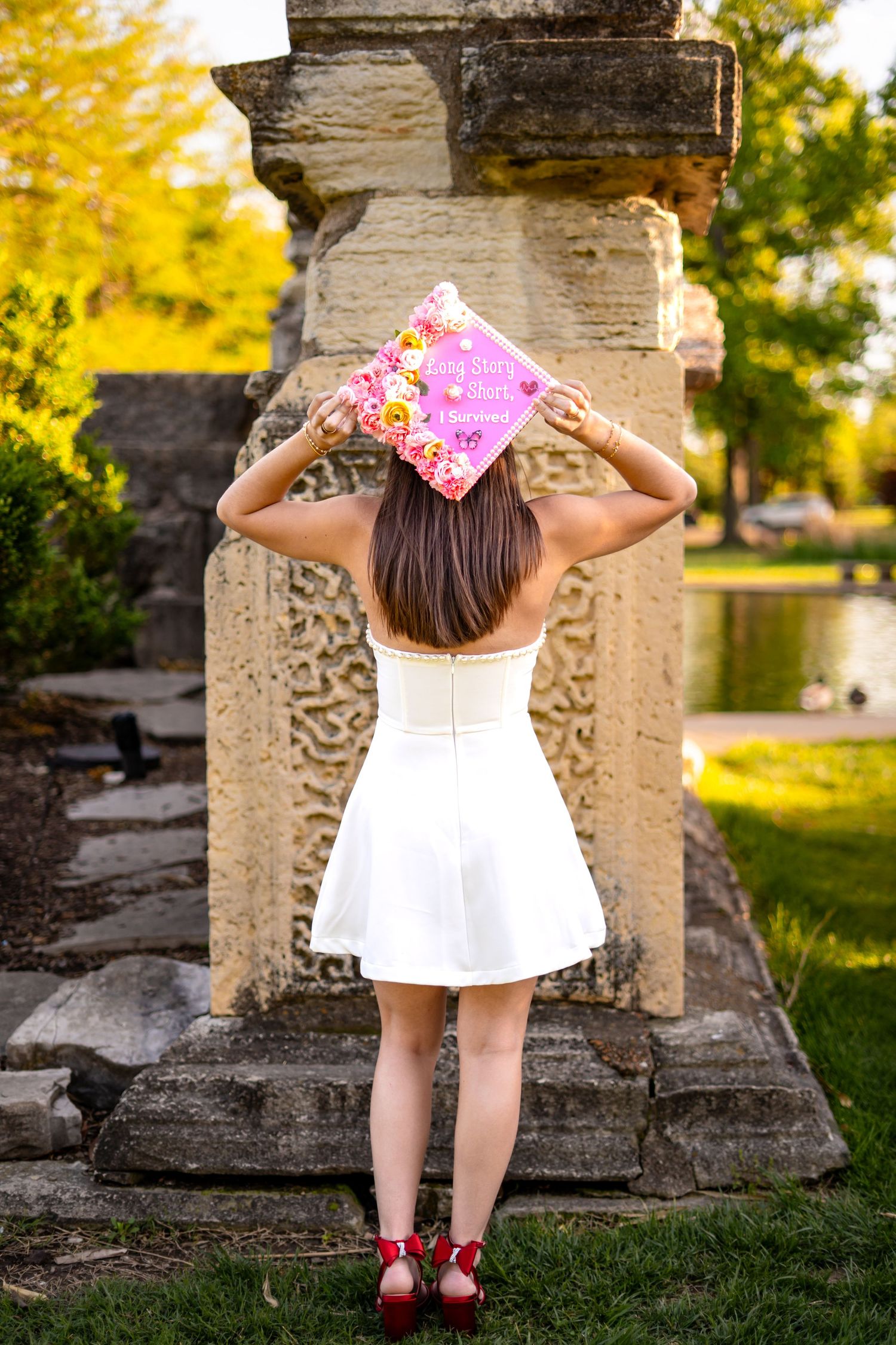 Graduate in white dress displays pink floral decorated graduation cap near stone columns by pond.