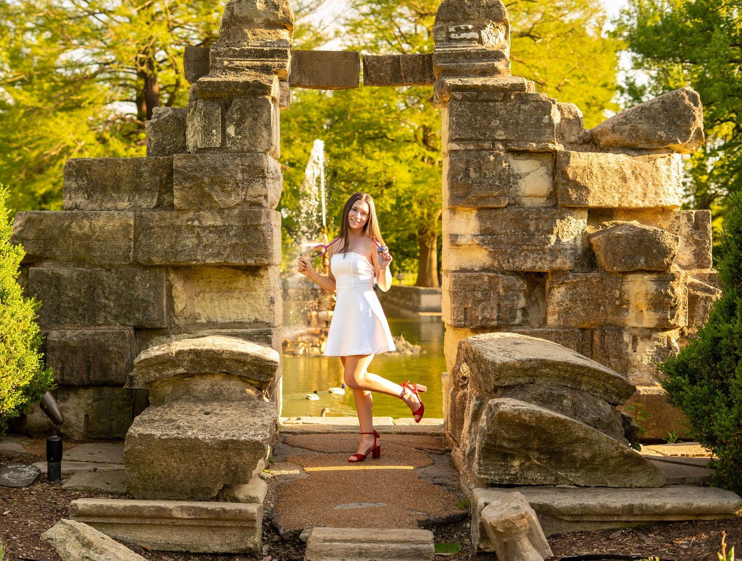 Nursing graduate in white dress poses in rustic stone archway with fountain and trees behind her.