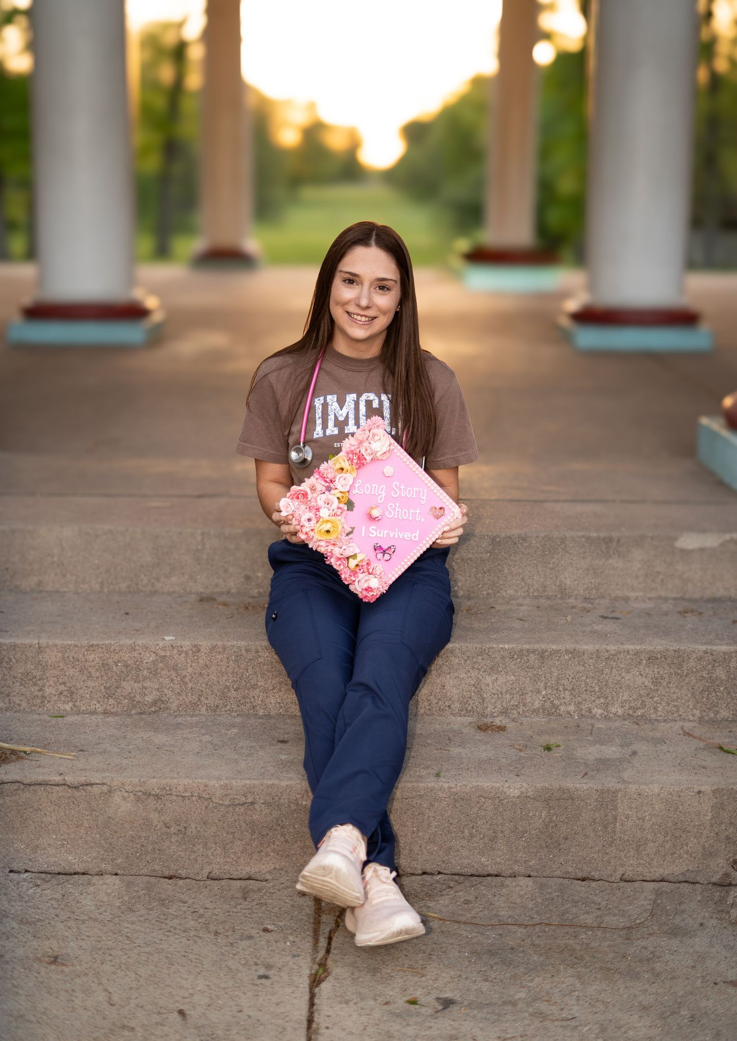 Nursing student in IMCU shirt sits on stone steps holding pink floral graduation cap at sunset.
