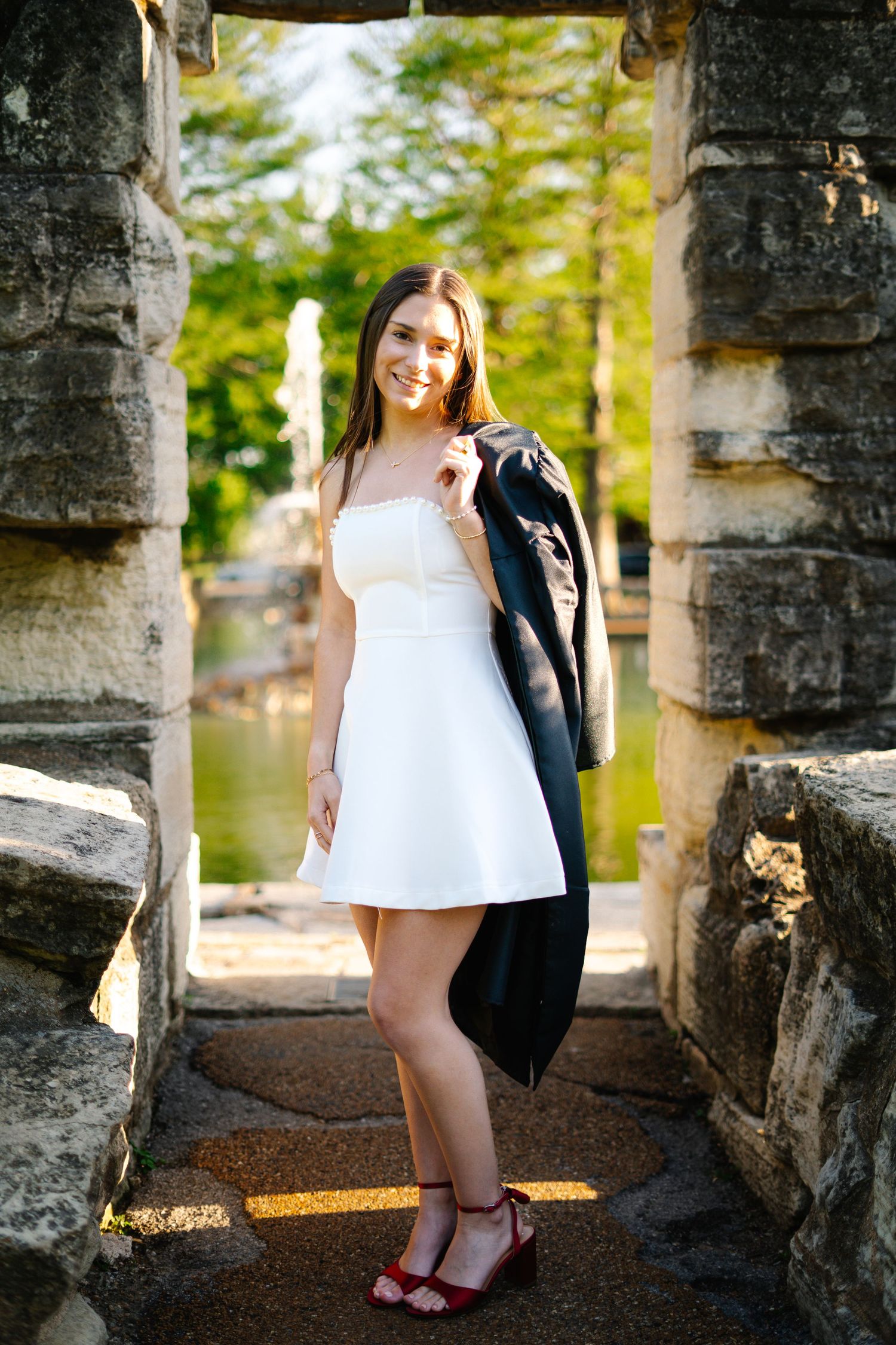 Smiling woman in white dress carries black graduation gown over shoulder while posing in stone archway near fountain.