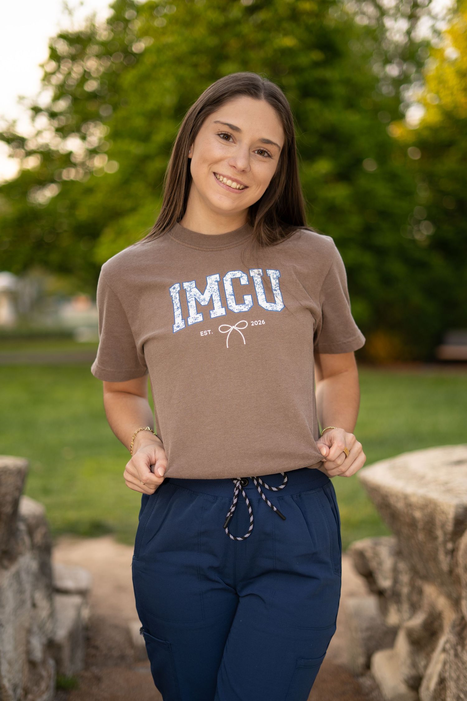 Smiling young woman in brown IMCU t-shirt and navy scrub pants poses outdoors in a park setting.