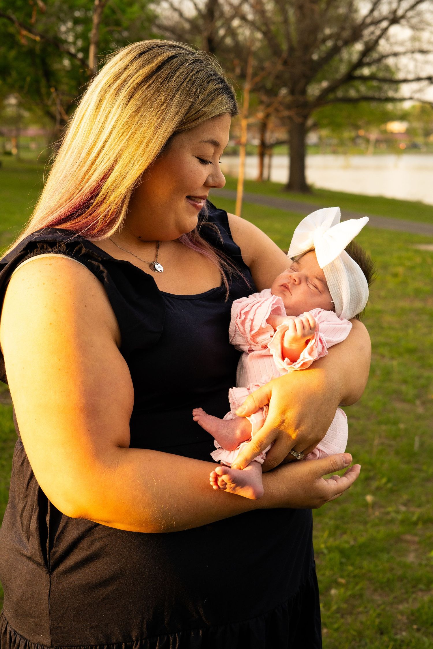 Woman in black dress tenderly holding newborn baby in pink outfit with white bow headband.