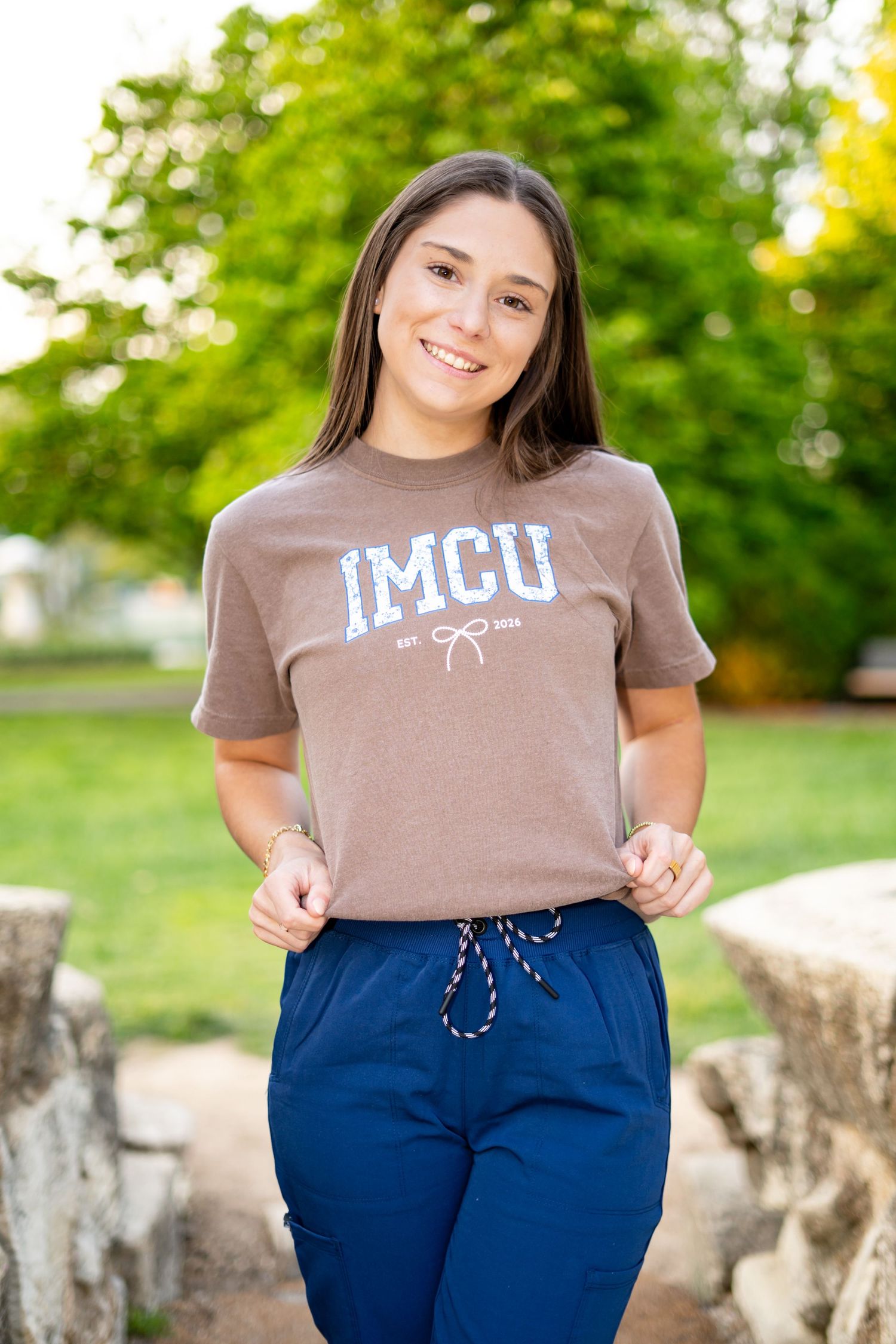 Young woman smiling and holding her IMCU brown t-shirt while wearing navy blue scrub pants outdoors.