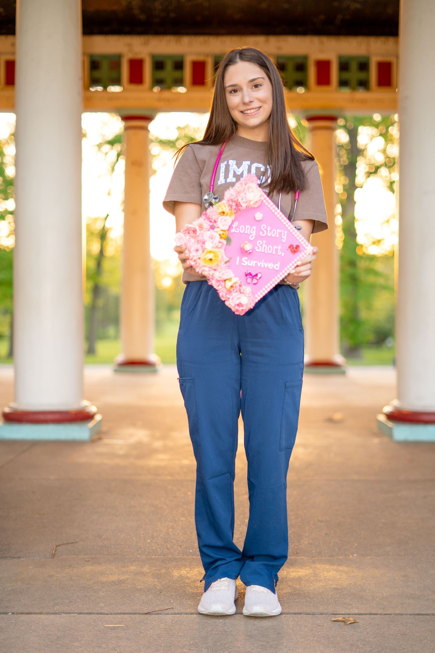 Nursing student in IMCU scrubs holds decorated pink graduation cap under pavilion columns.