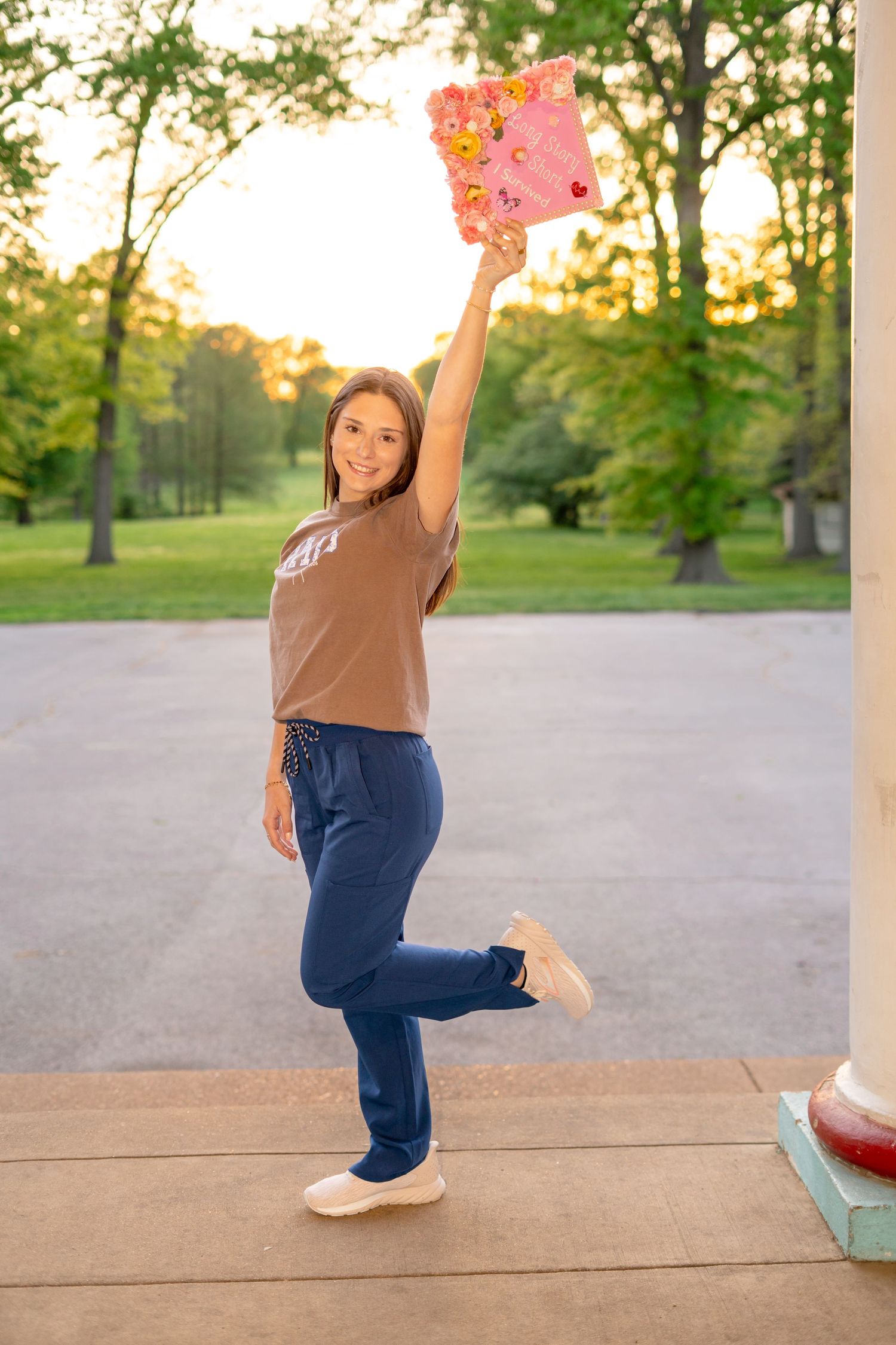 Nursing graduate in scrubs joyfully raises decorated pink graduation cap outdoors at sunset.