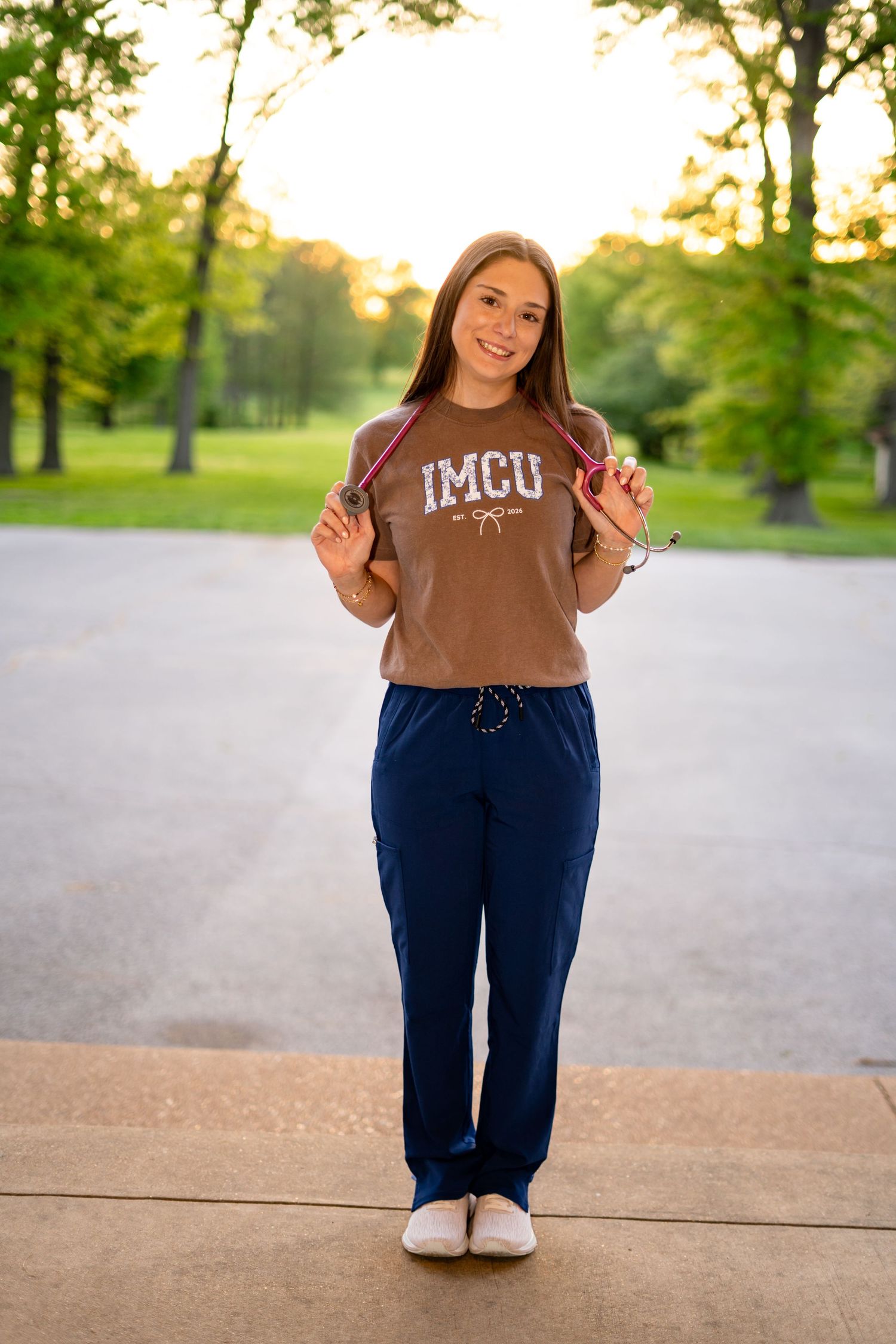 Young woman in IMCU scrub top and navy pants holds stethoscope at golden hour.