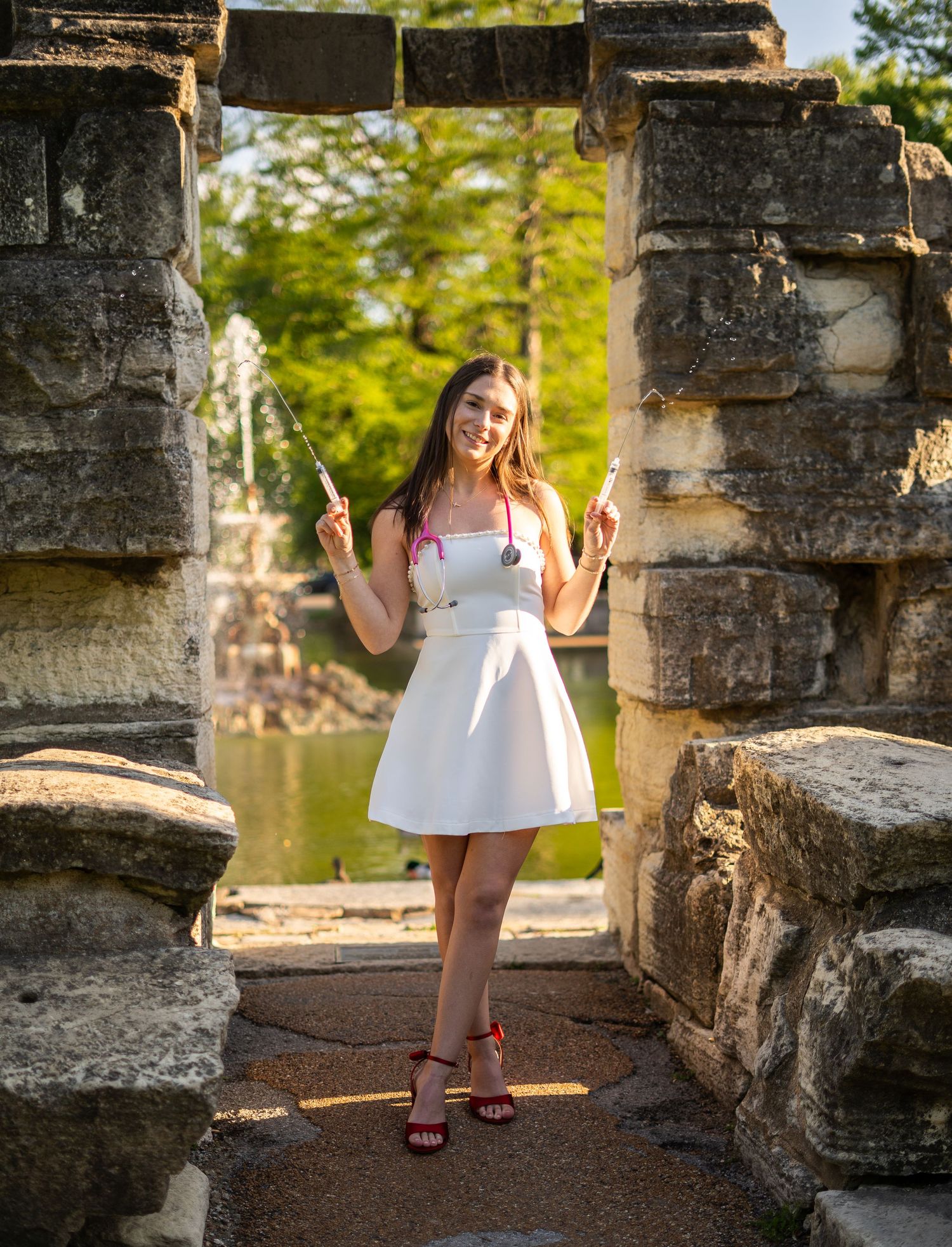 Nursing graduate in white dress holds syringes with pink stethoscope posing between stone columns near garden fountain.