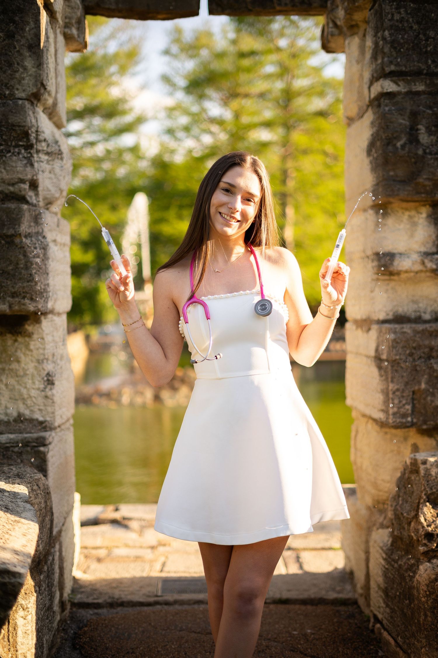 Smiling nursing graduate in white dress holds syringes with pink stethoscope at stone arch outdoors.