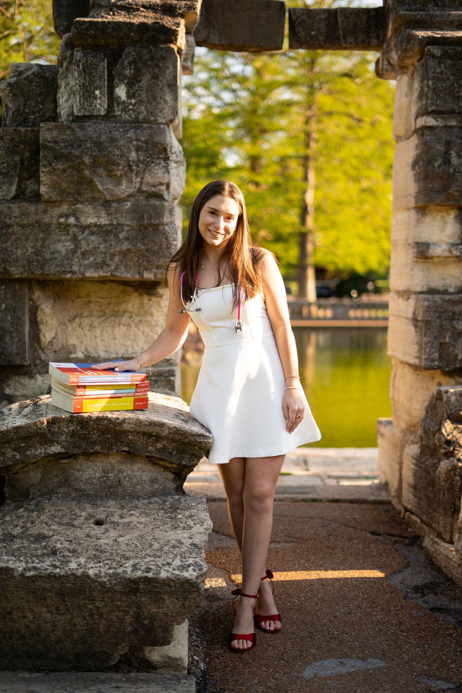 Young woman in white dress poses with textbooks at stone ruins near a pond.