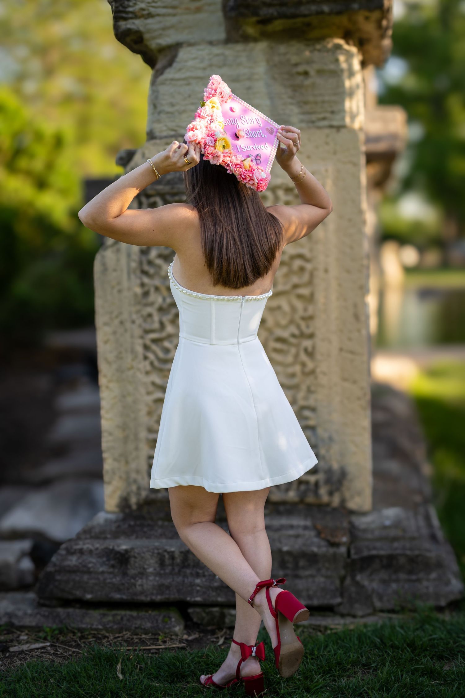 Nursing graduate in white dress and red heels places pink floral cap on head near stone columns.