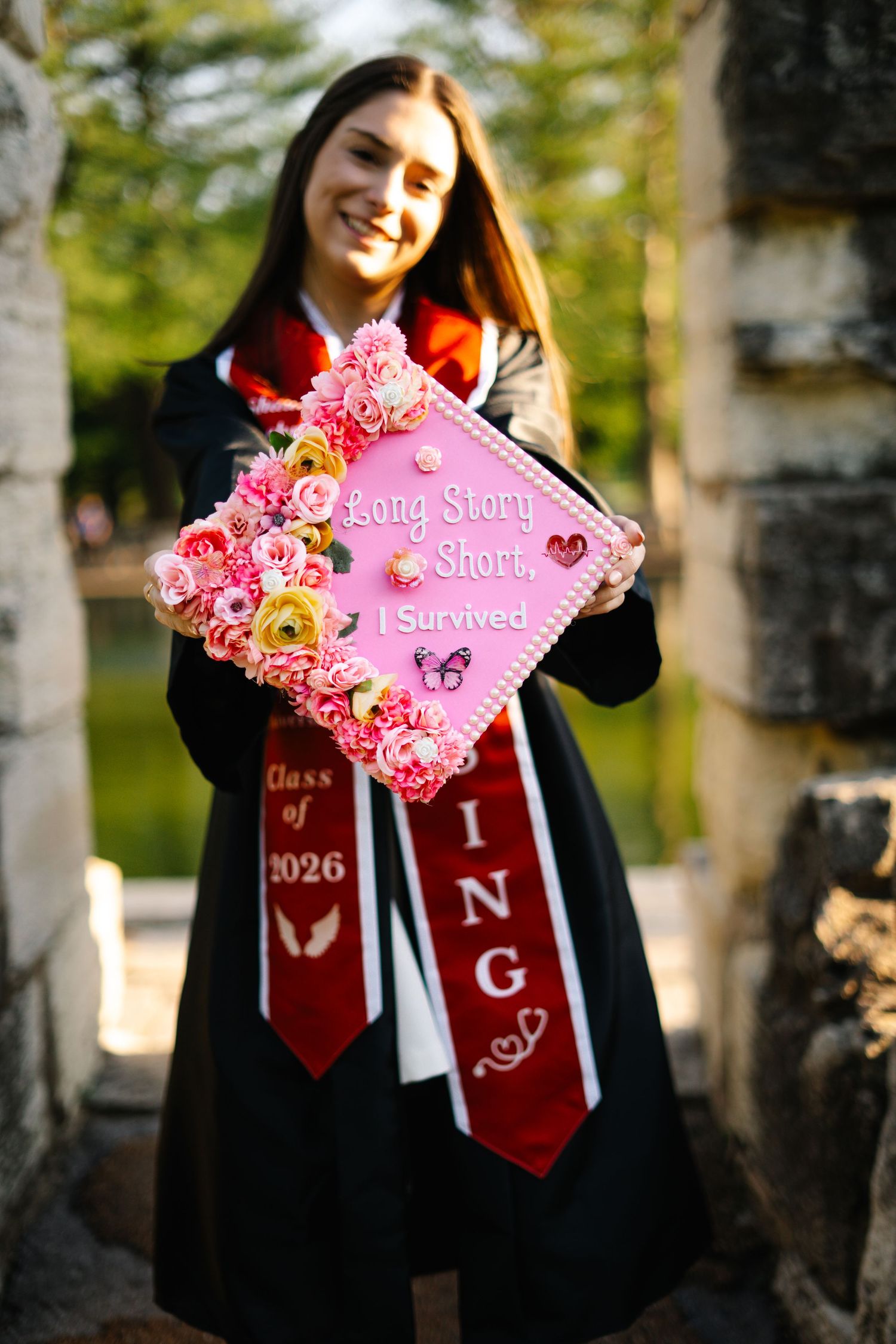 Smiling nursing graduate holds decorated pink cap reading Long Story Short I Survived with floral accents and red stole.