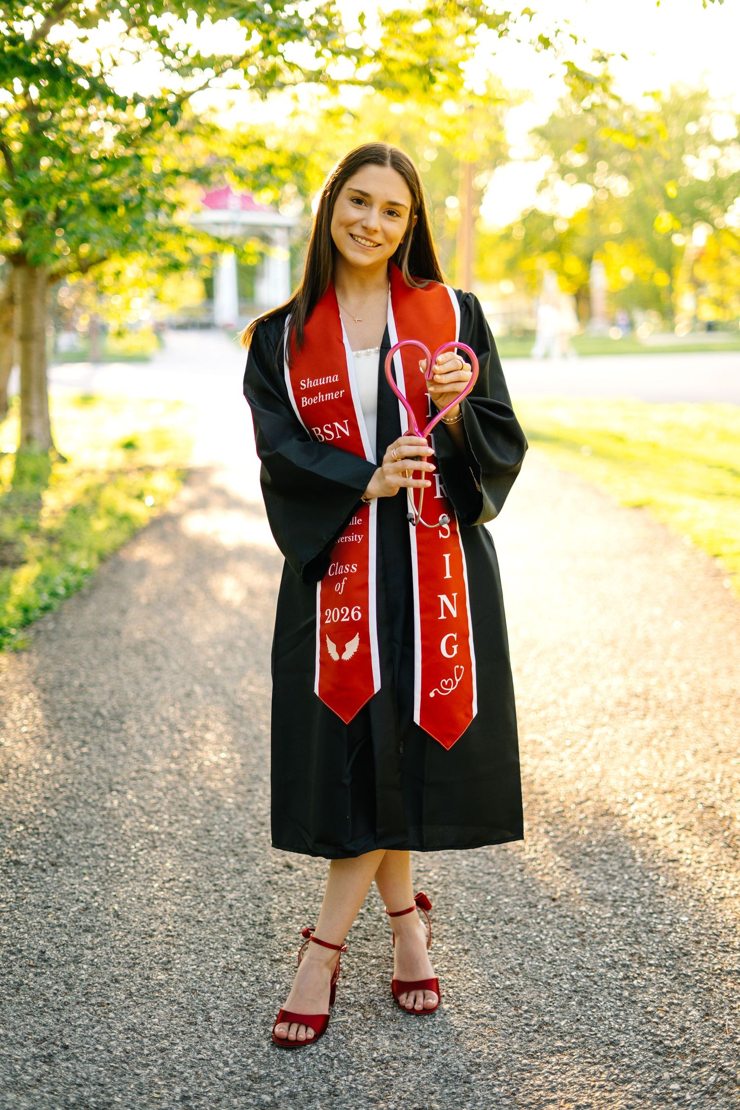 Nursing student in black graduation gown with red BSN stole smiles while standing on tree-lined campus path.
