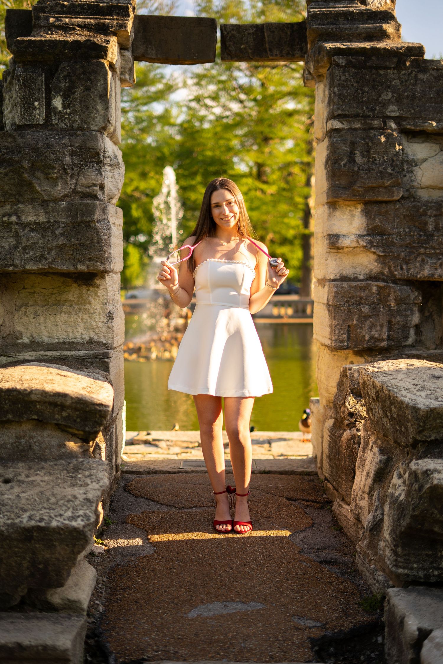 Smiling nursing graduate in white dress stands in stone archway holding items near sunlit fountain.