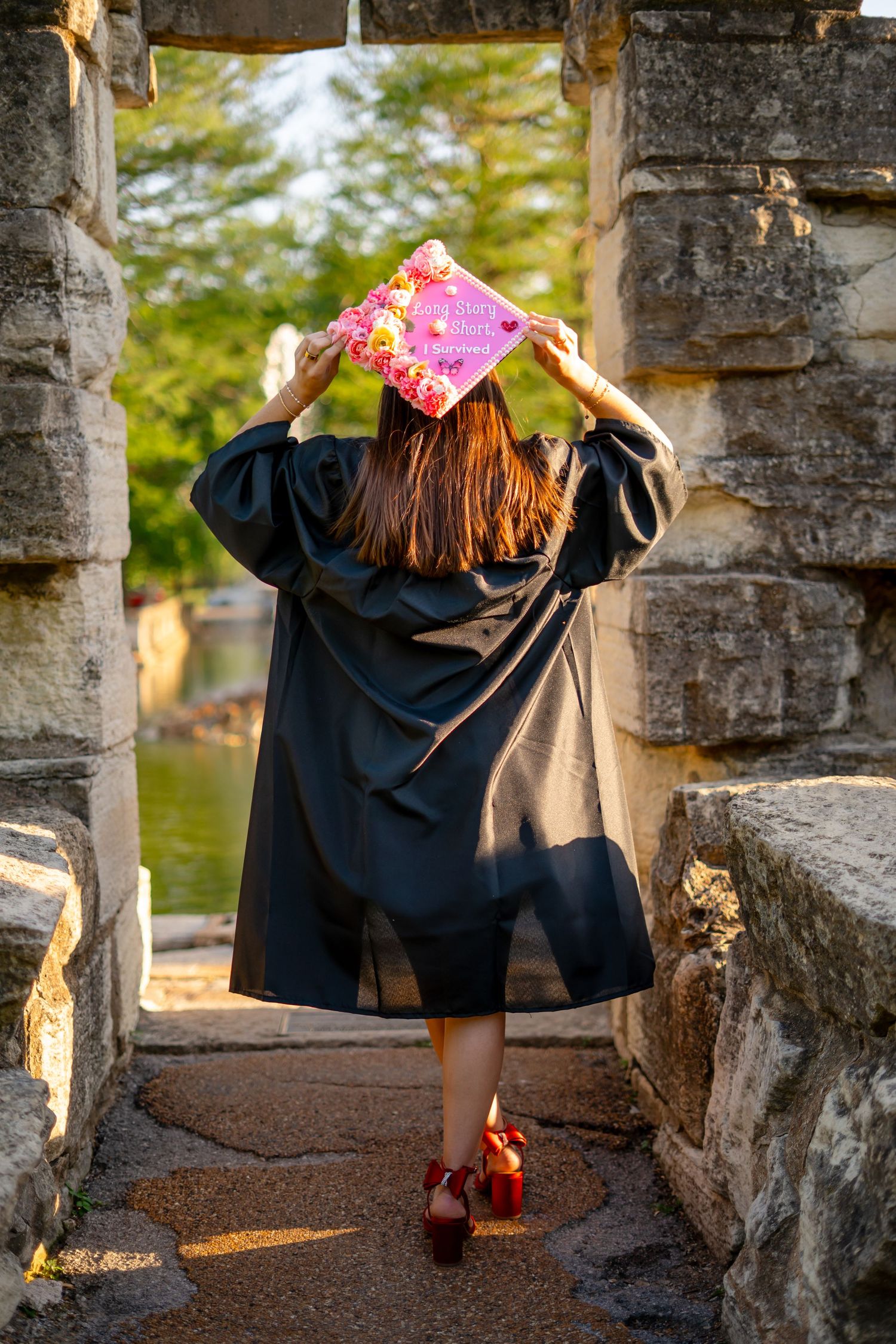Nursing graduate in black gown and red heels walks away holding pink decorated graduation cap between stone pillars.