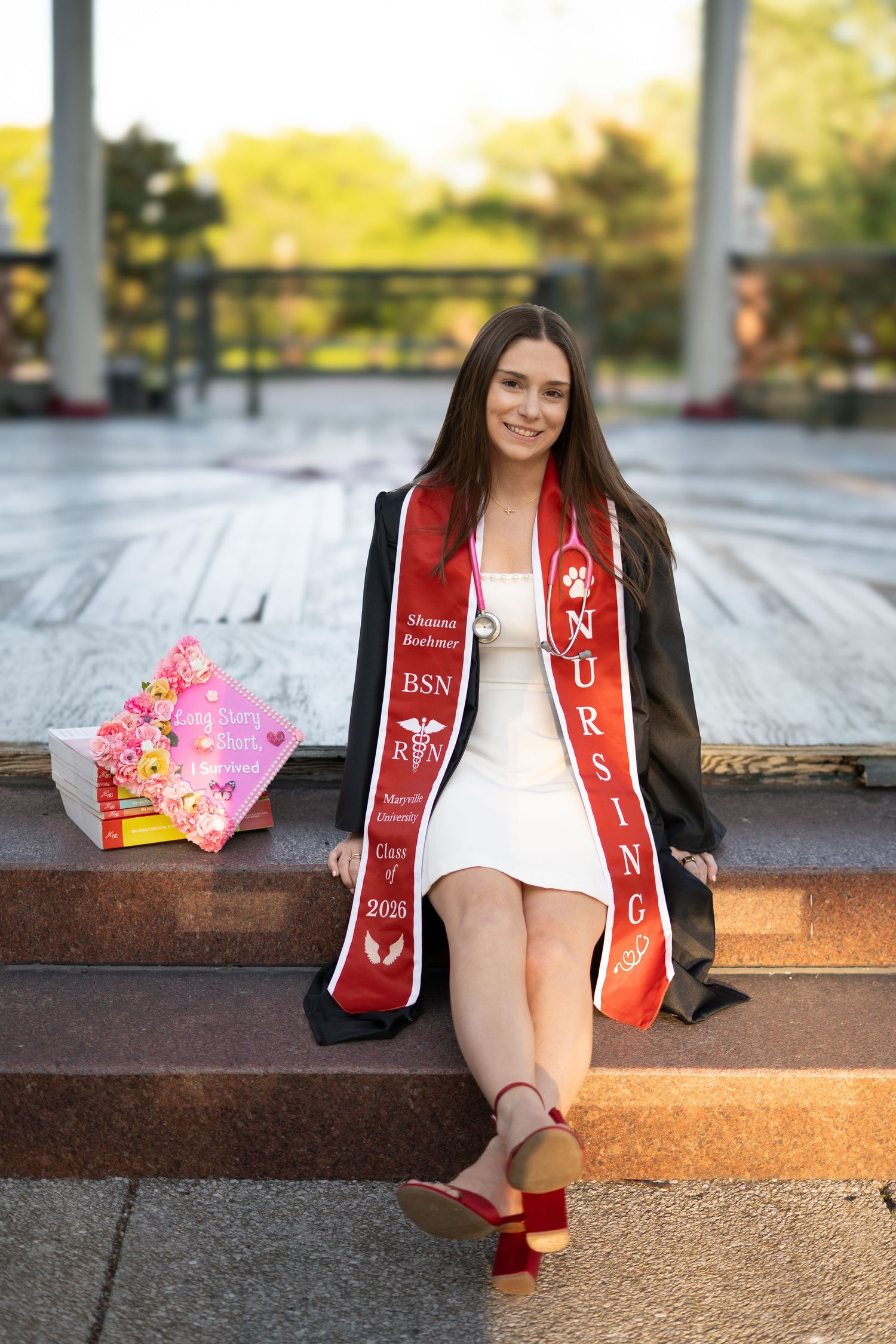 Nursing graduate in gown and red stole sits on steps holding floral decorated cap and bouquet outdoors.