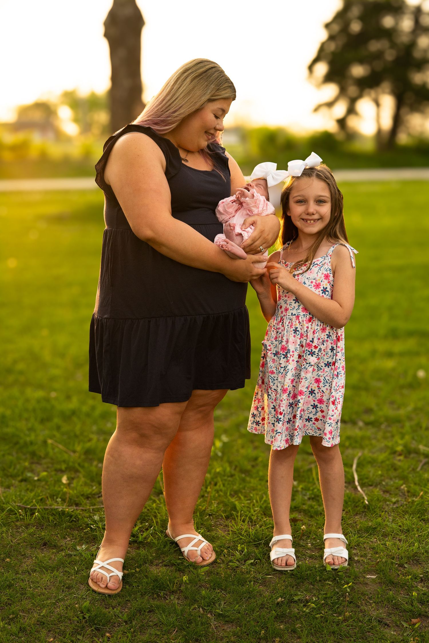 Blonde mother in black dress holding newborn baby beside smiling young daughter in floral dress at sunset park.