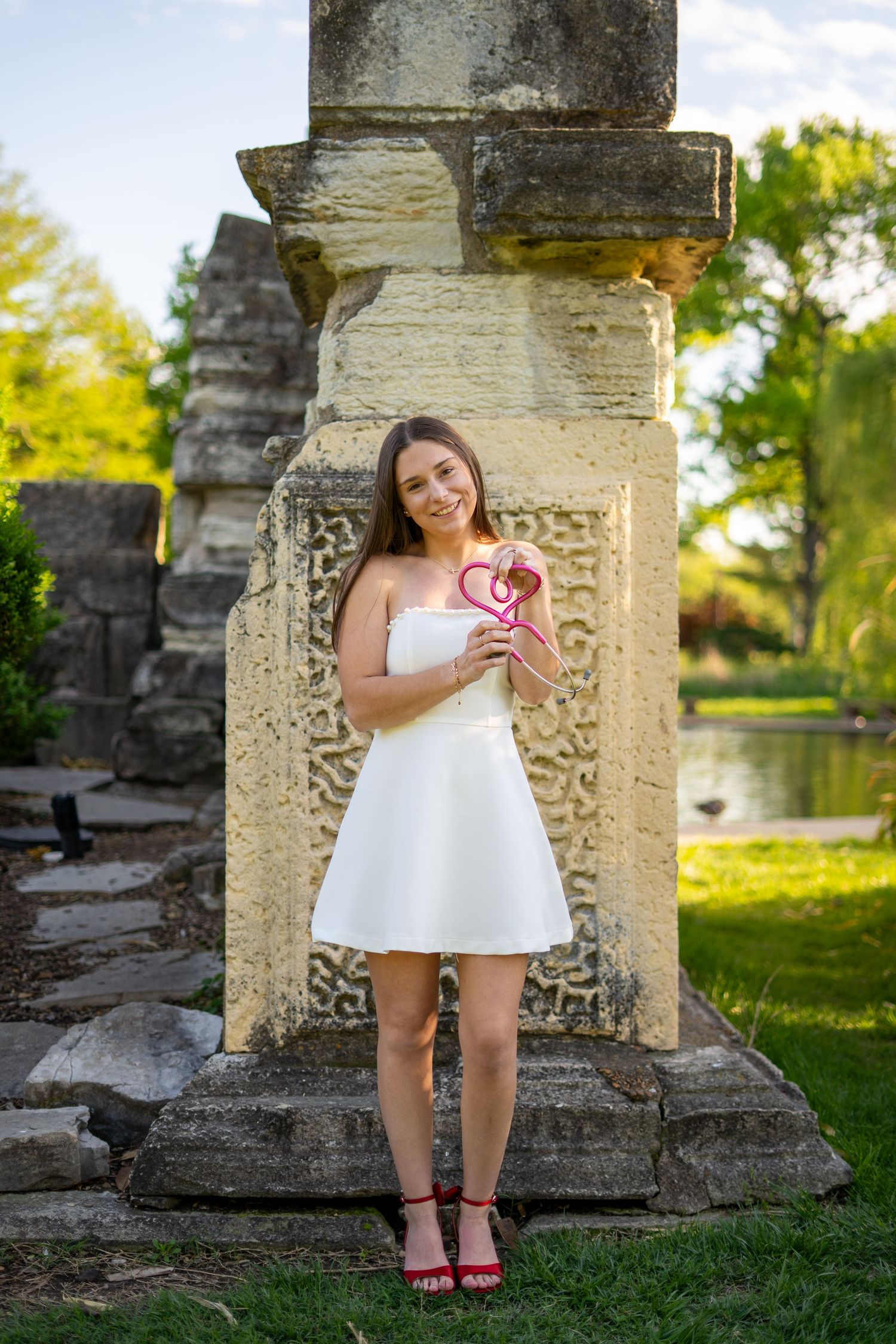Smiling young woman in white strapless dress holds pink heart prop beside ornate stone column in park.