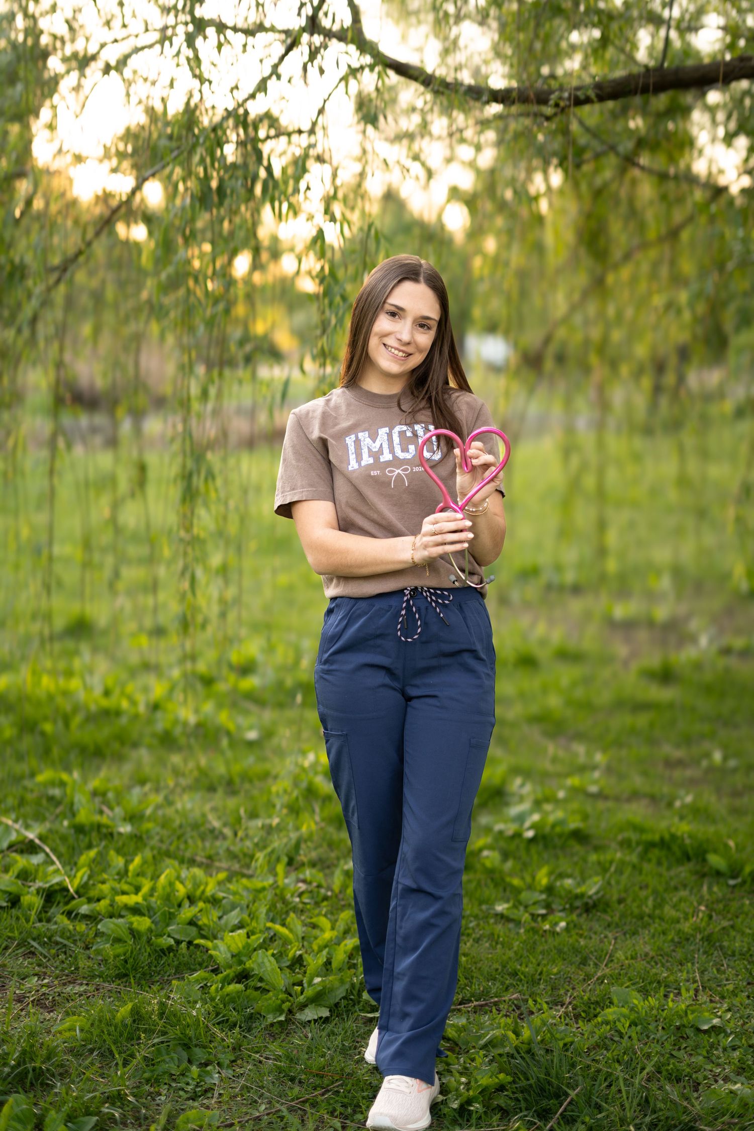 Young woman in IMCO shirt and navy pants smiling while holding a heart-shaped wand in a lush green park.