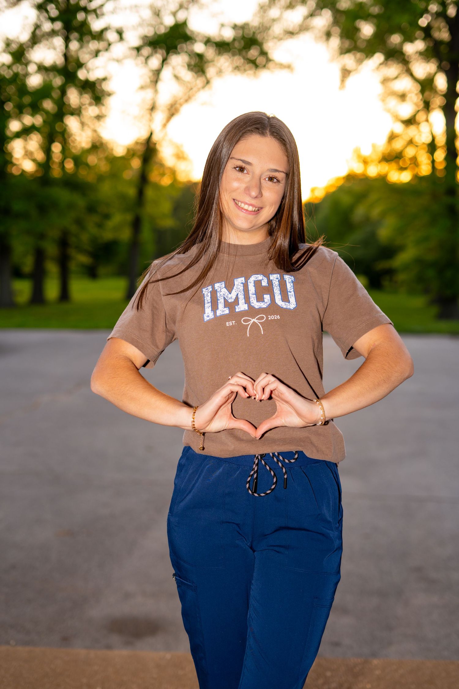 Young woman in IMCU brown t-shirt and blue scrub pants making a heart shape with her hands outdoors at sunset.