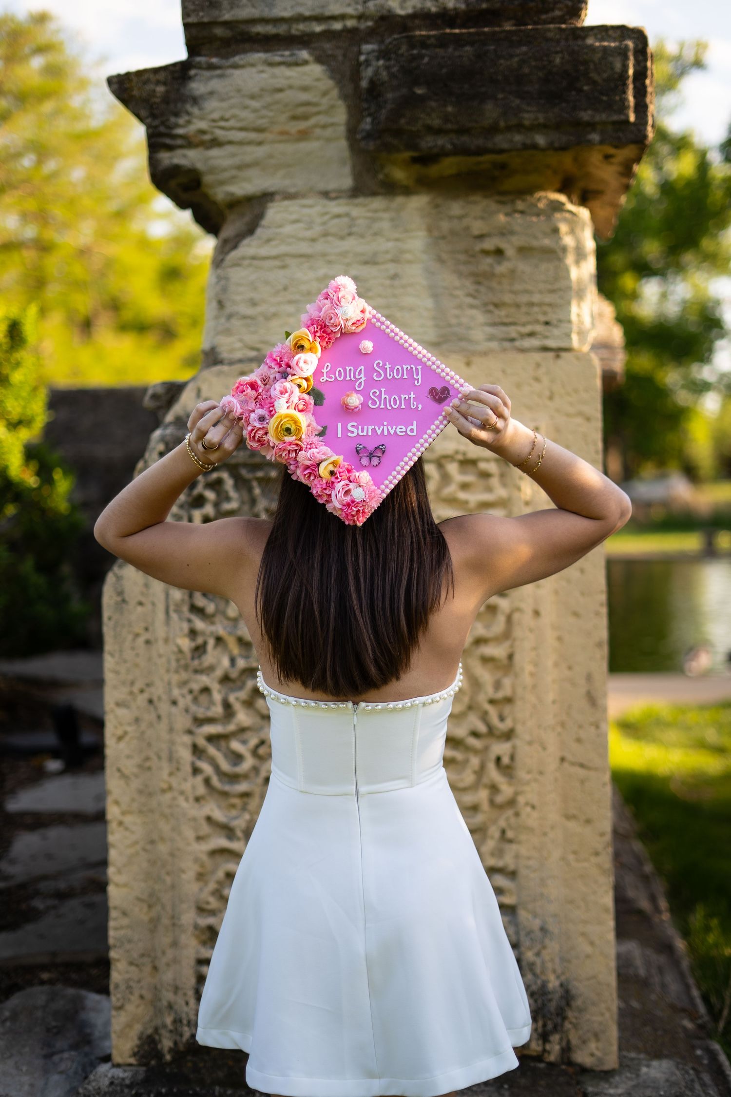 Graduate holds decorated purple floral cap reading 'Long Story Short, I Survived' wearing white dress outdoors.