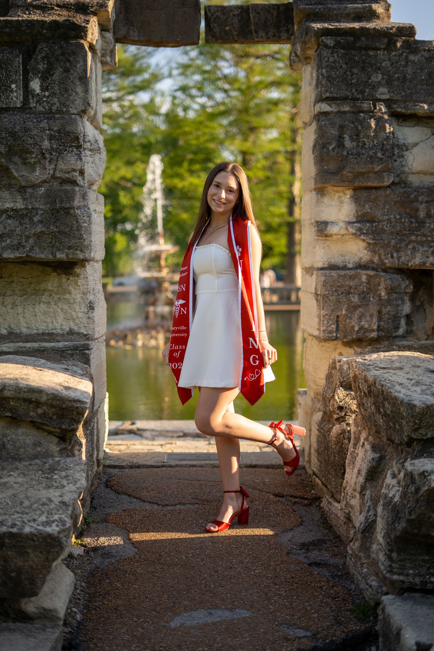 Nursing graduate in white dress and red heels poses playfully in stone archway with red graduation stole.