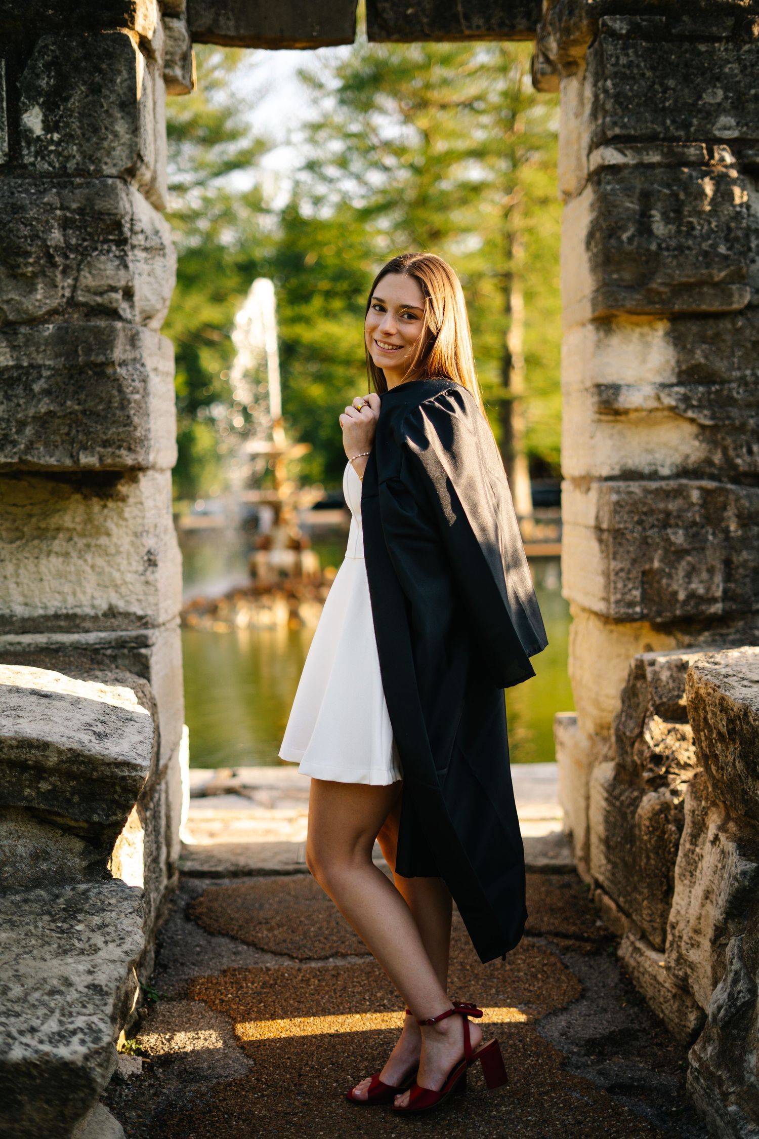 Smiling young woman in white dress drapes black graduation gown over shoulder between stone archway near fountain.