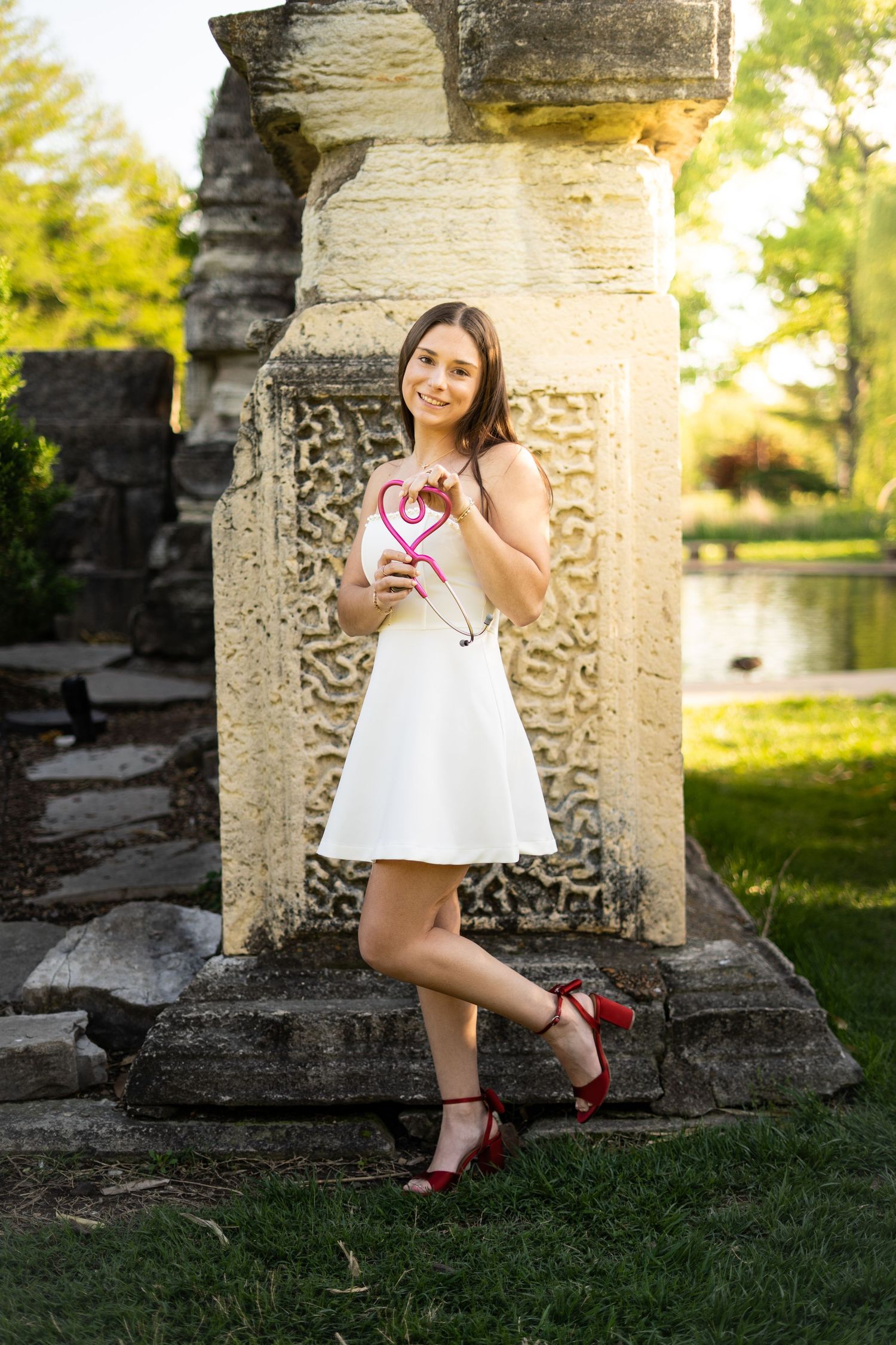 Young woman in white dress and red heels poses with pink heart beside carved stone pillar at sunset.