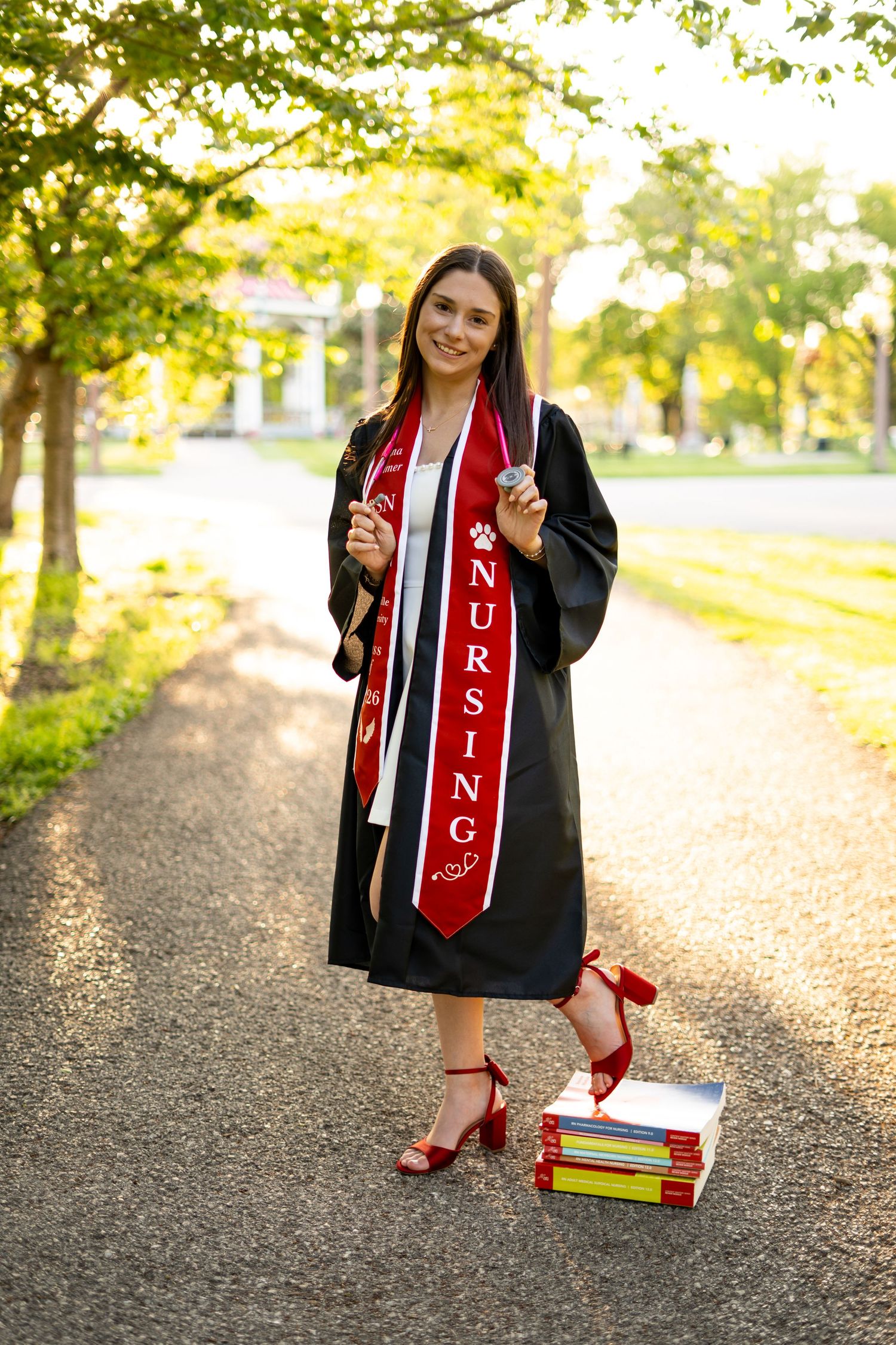 Nursing graduate in black cap and gown with red sash posing on a tree-lined campus pathway with textbooks.