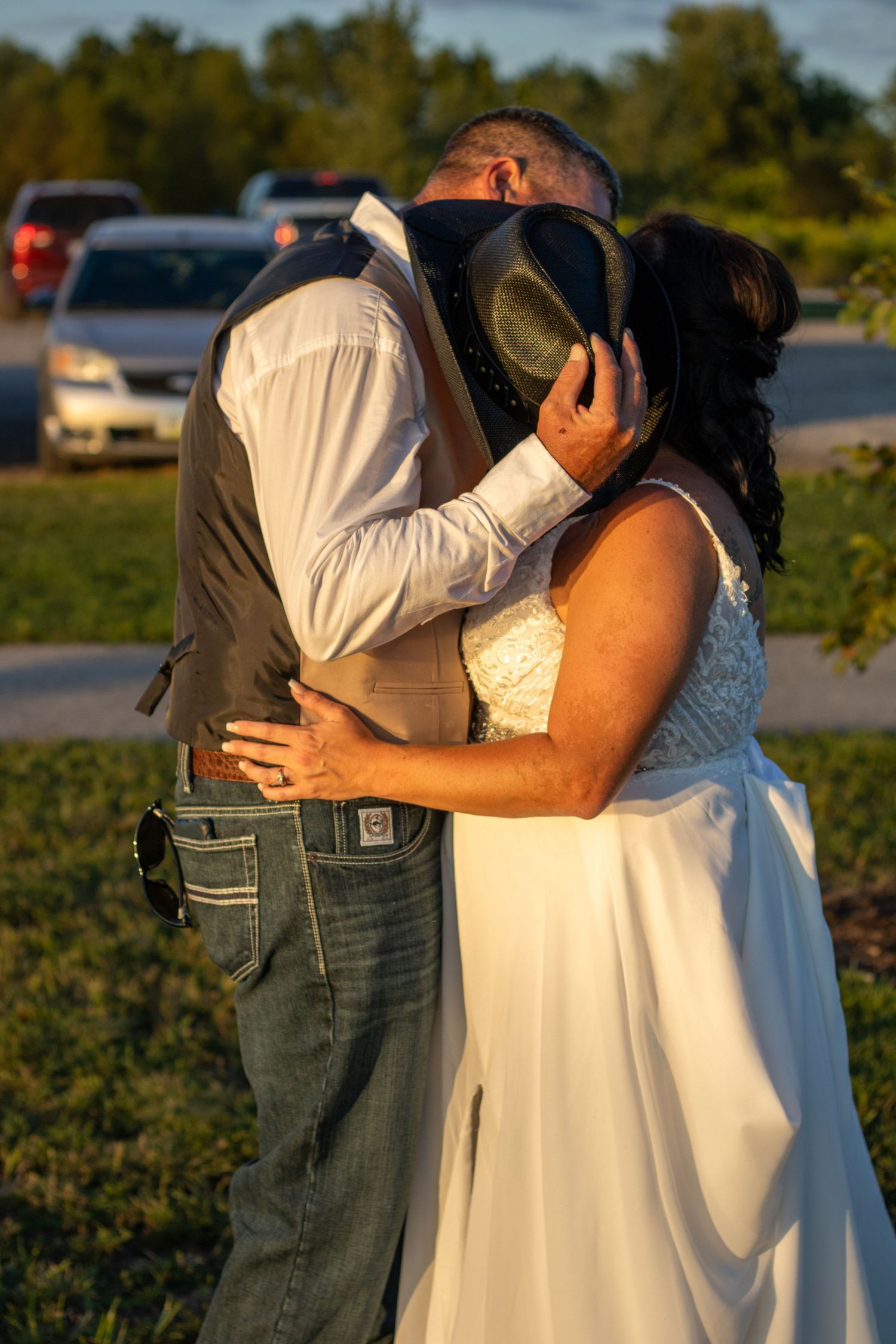 Bride and groom sharing a kiss hidden behind a cowboy hat outdoors.