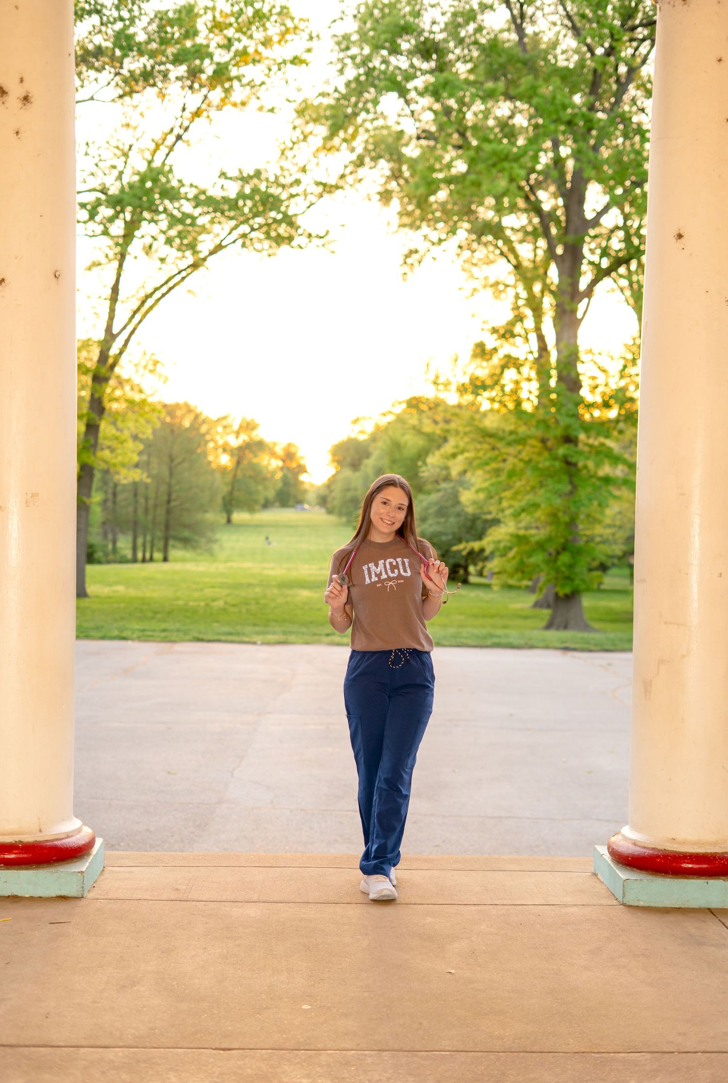 Young woman in IMCU nursing scrubs walks through white columns at sunset in park.