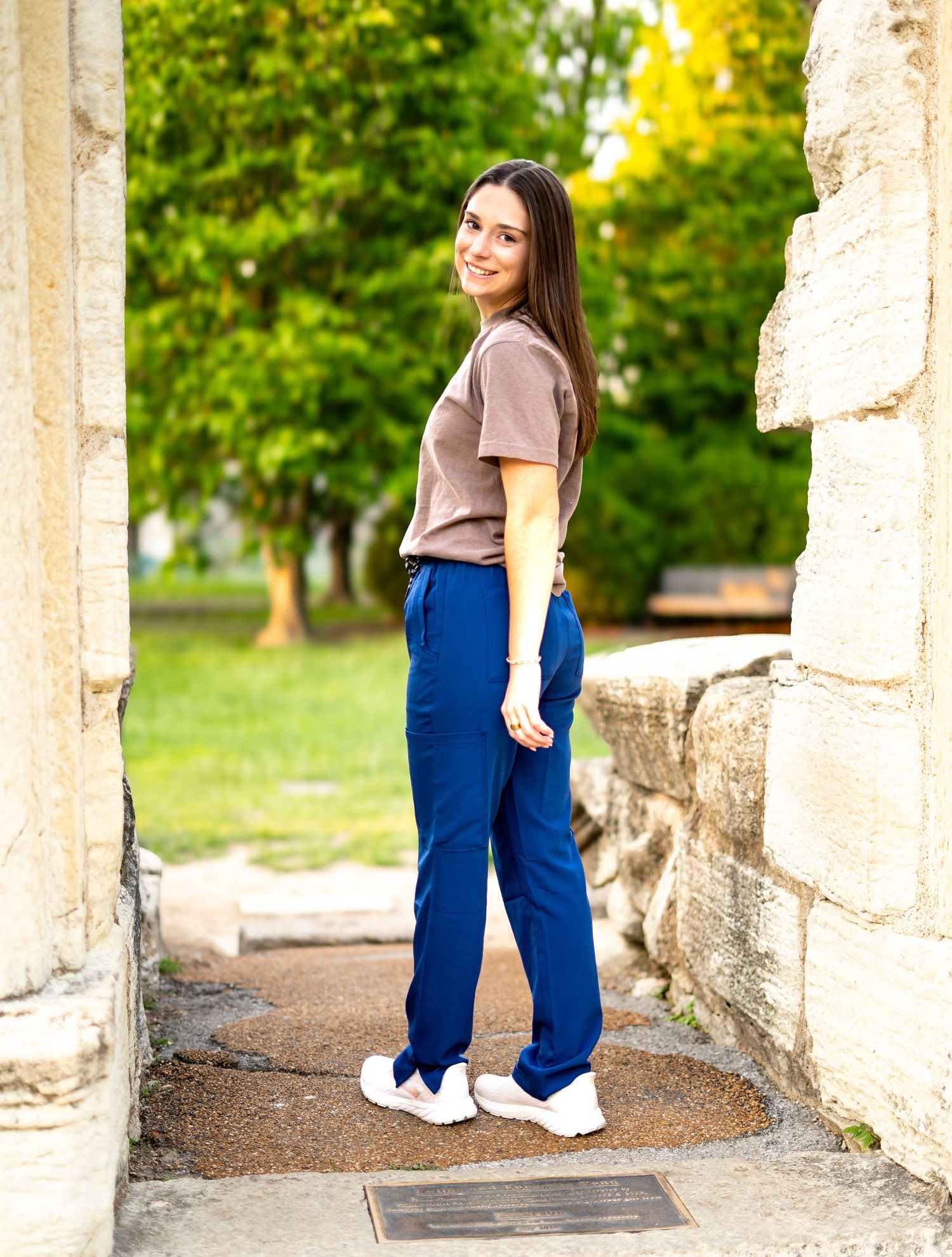 Young woman in nursing scrubs poses sideways between stone columns in outdoor setting.
