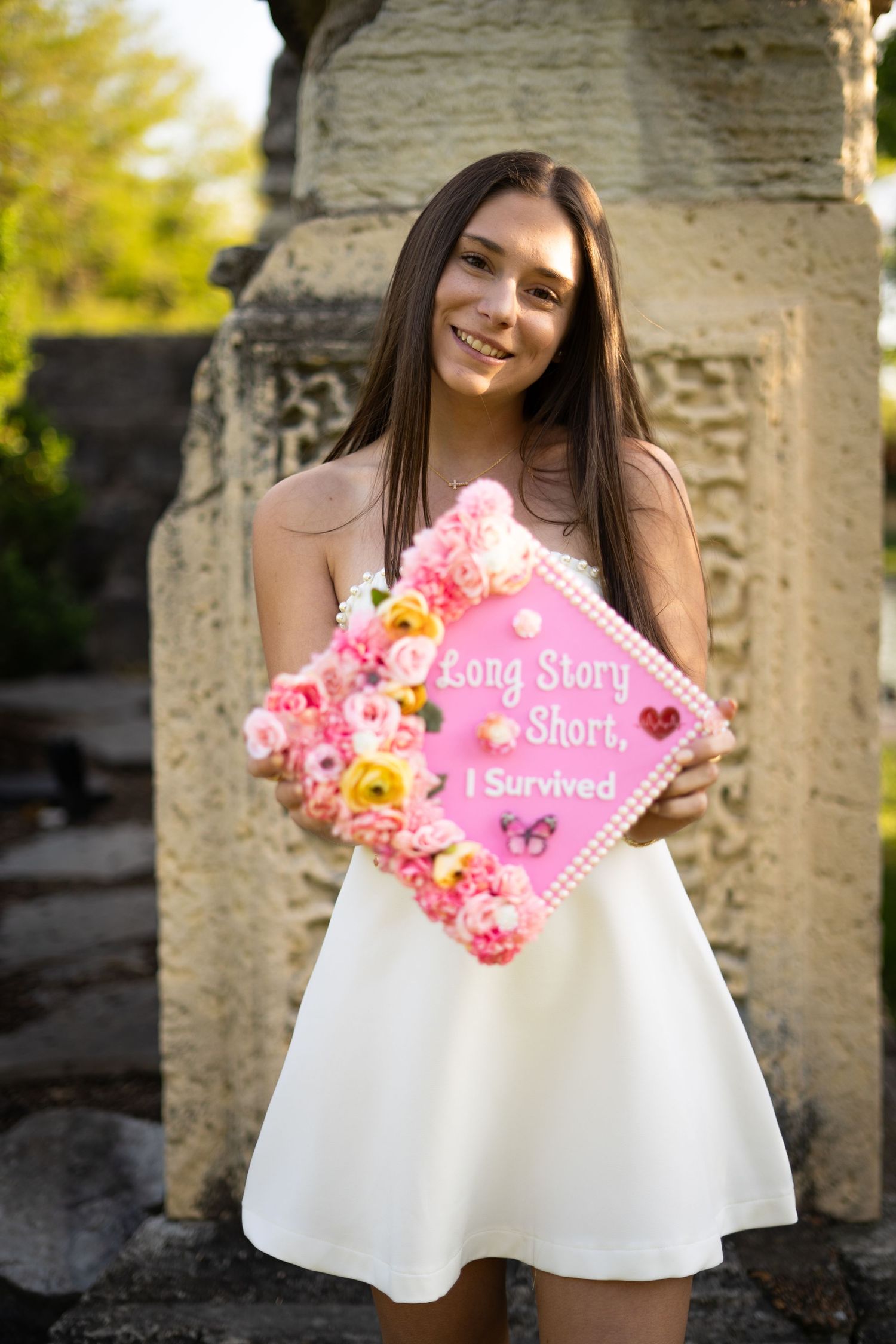 Smiling graduate holds pink floral cap reading Long Story Short I Survived beside stone column in park.