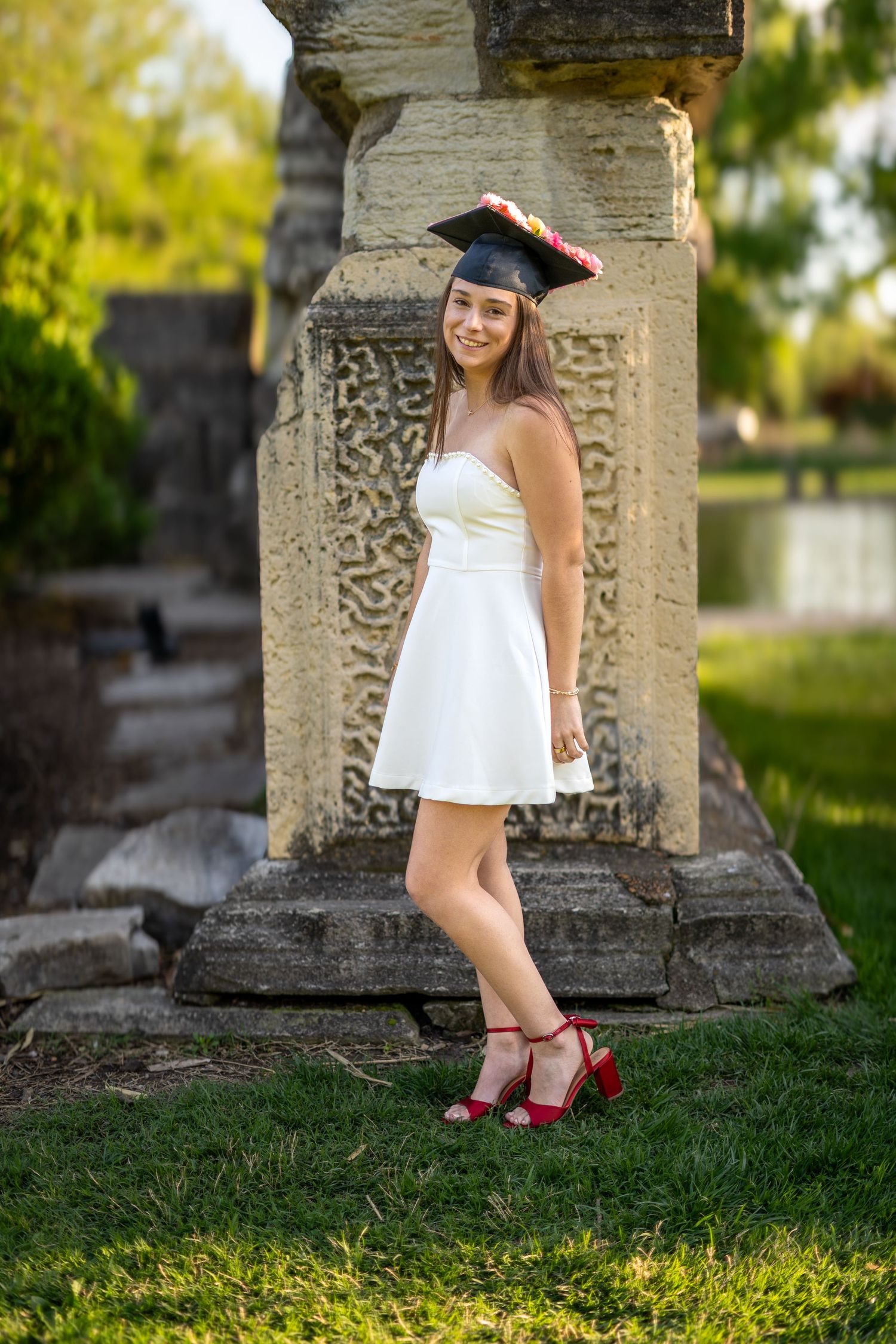Smiling nursing graduate in white dress and cap poses beside ornate stone column in garden.