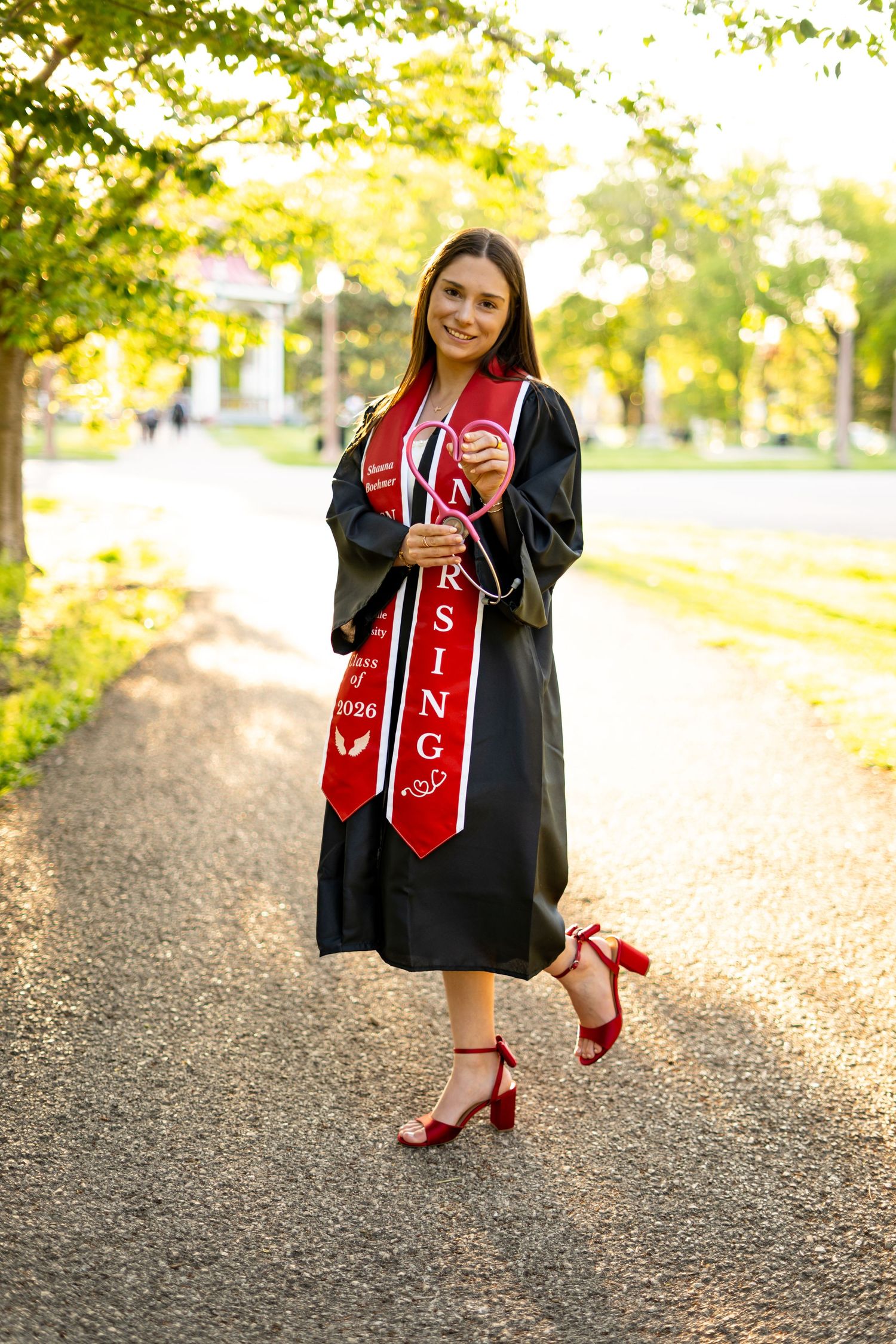 Happy nursing graduate in black gown with red NURSING stole poses on sunlit campus pathway in red heels.