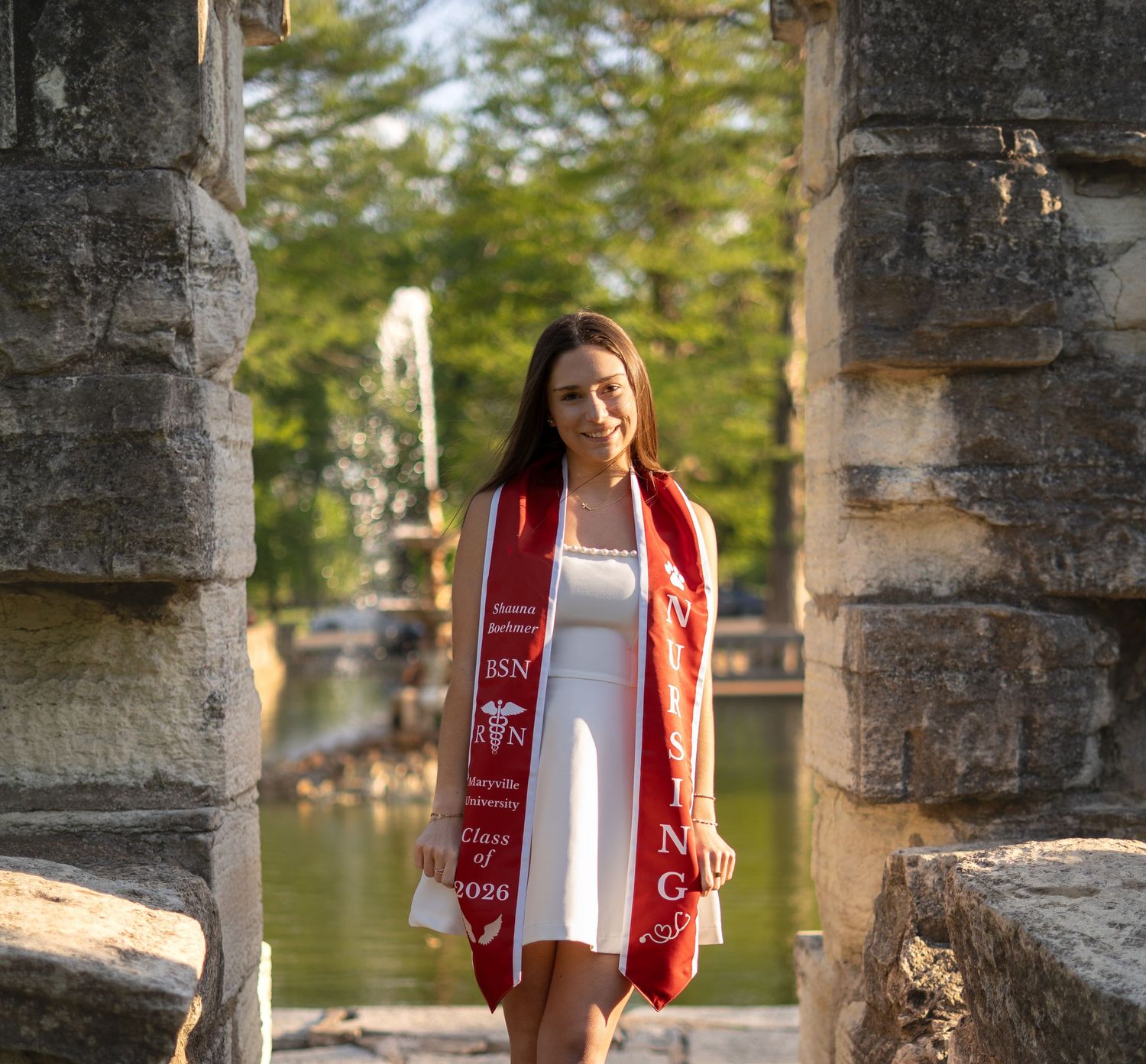 Nursing graduate wears red BSN stole and white dress standing in stone archway near pond.