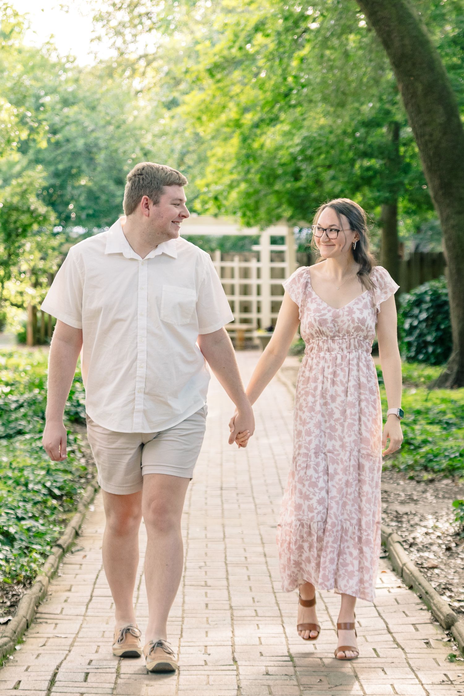 Couple walks hand-in-hand on brick garden path, smiling at each other in sunlit park.