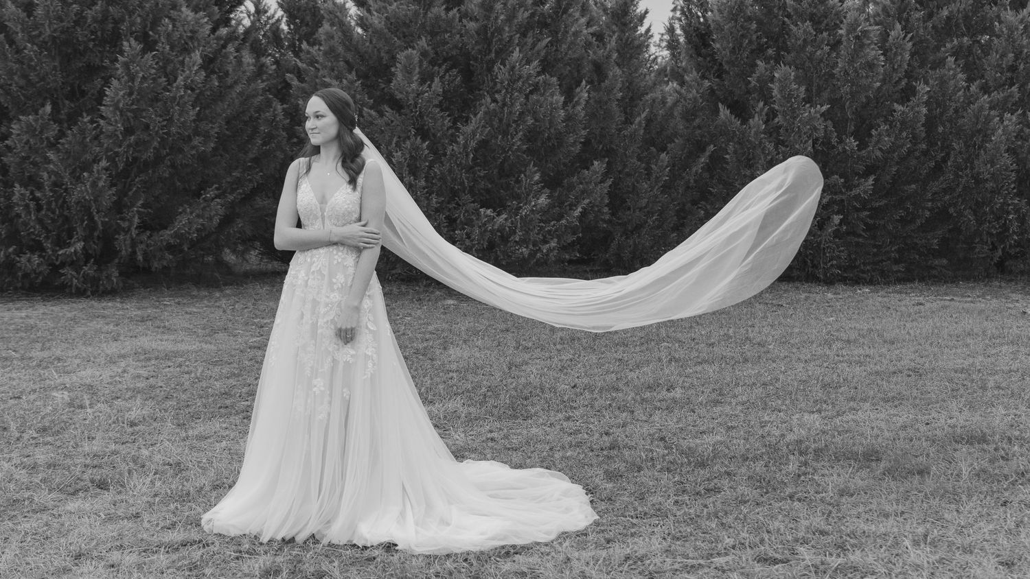 A black and white photo of a bride in a flowing gown with a long veil billowing in the wind outdoors.
