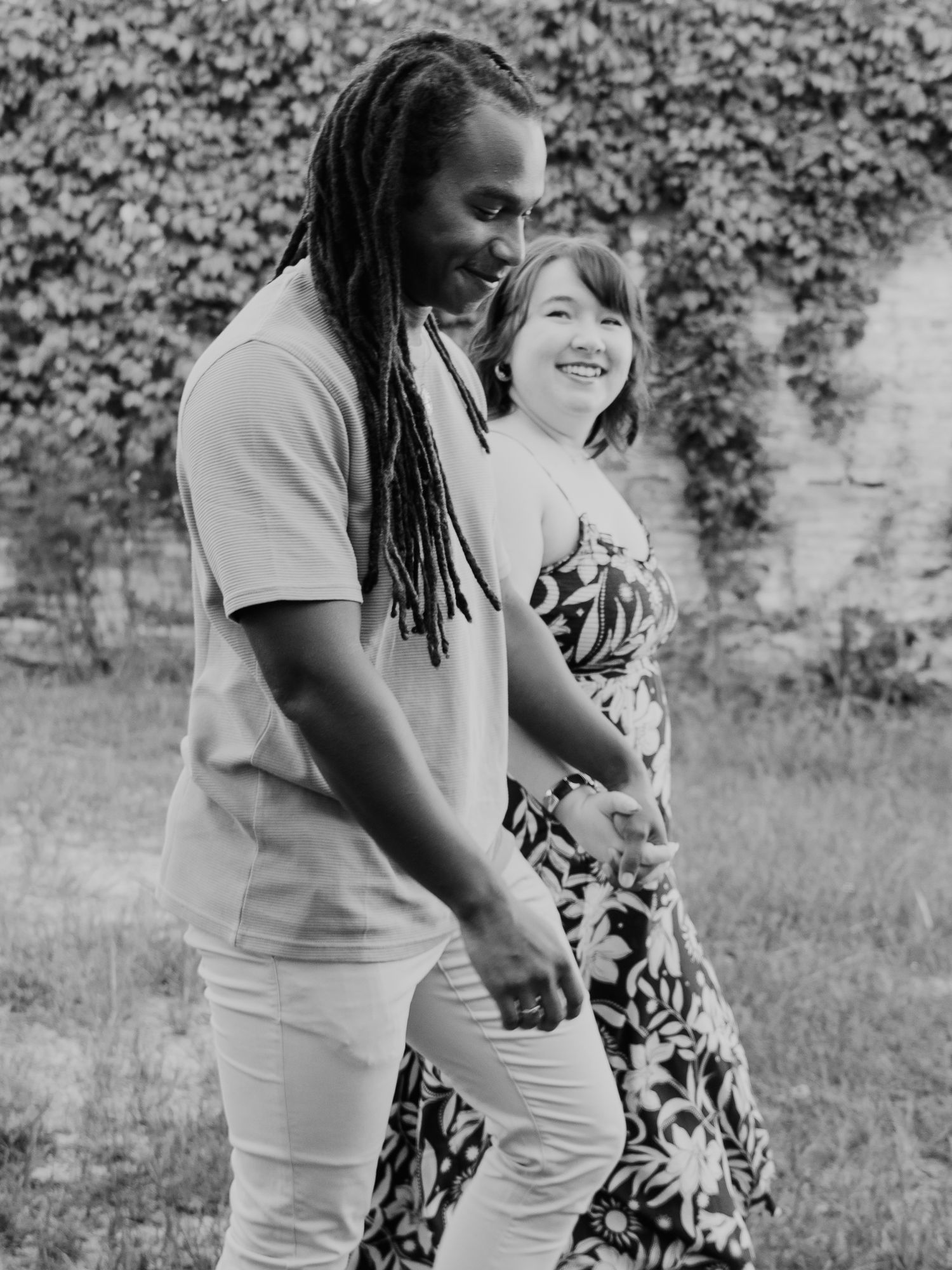 Black and white photo of a smiling couple walking hand in hand outdoors near an ivy-covered wall.