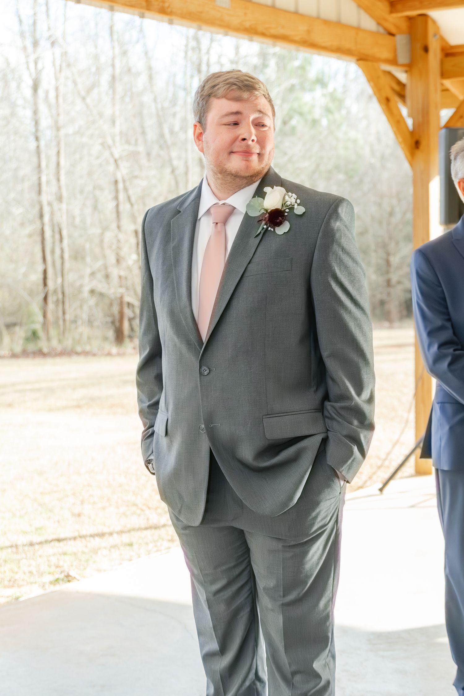 A groomsman in a gray suit with pink tie and boutonniere stands under a wooden pavilion during a wedding ceremony.