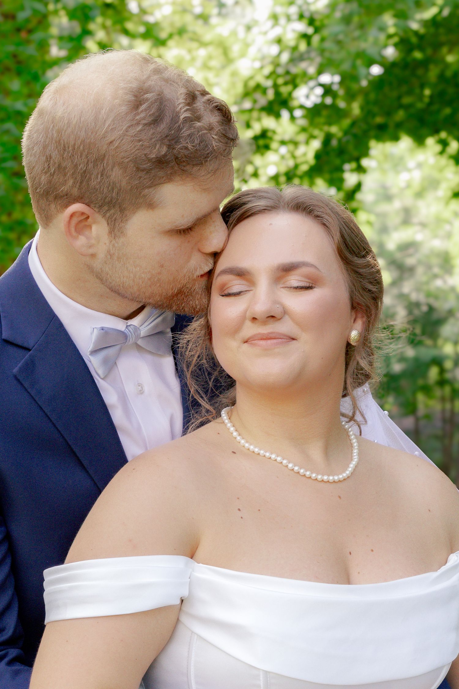 Groom in blue suit kissing bride in white off-shoulder gown on cheek outdoors.