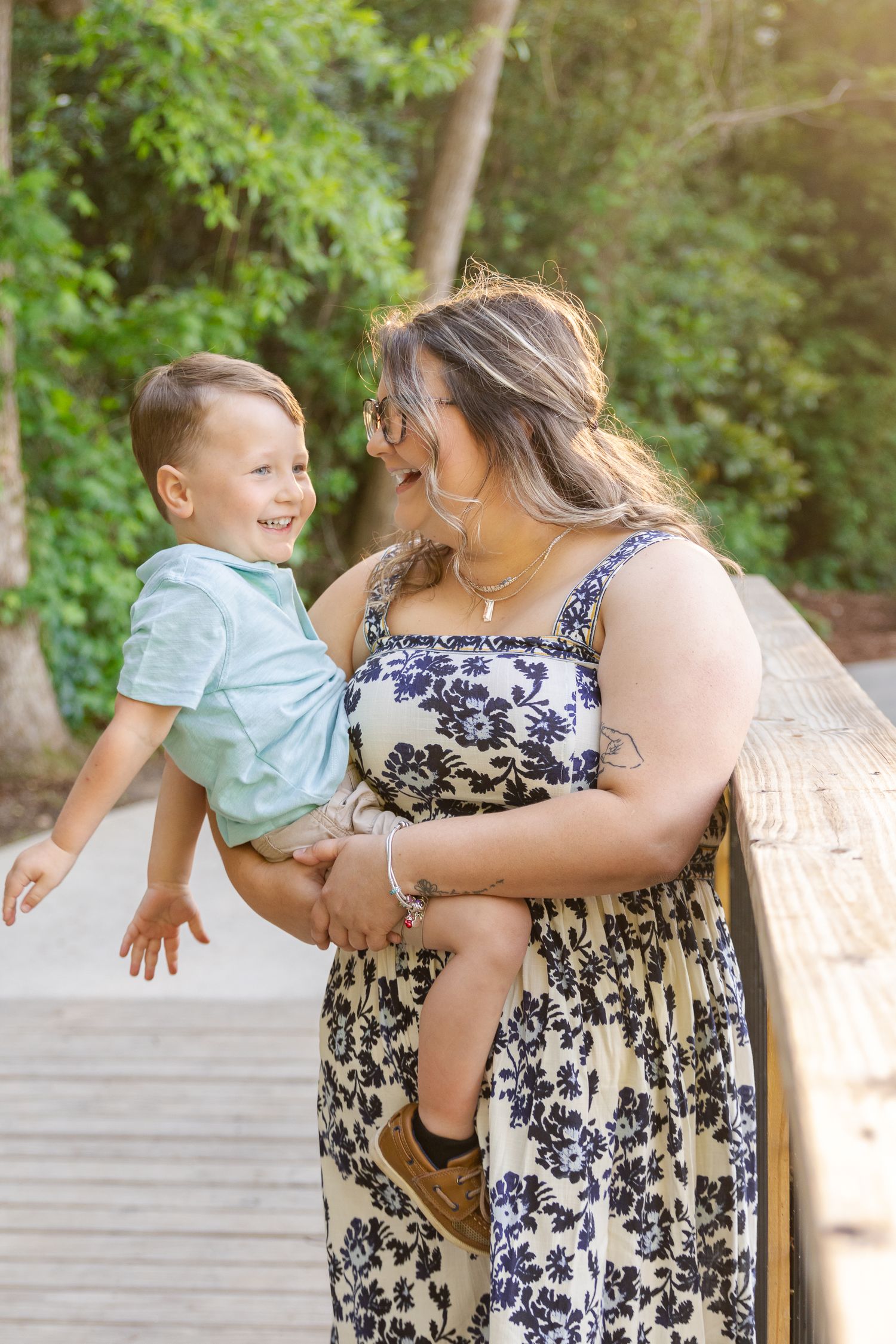 Joyful mother holding her laughing toddler son on a wooden bridge surrounded by greenery.