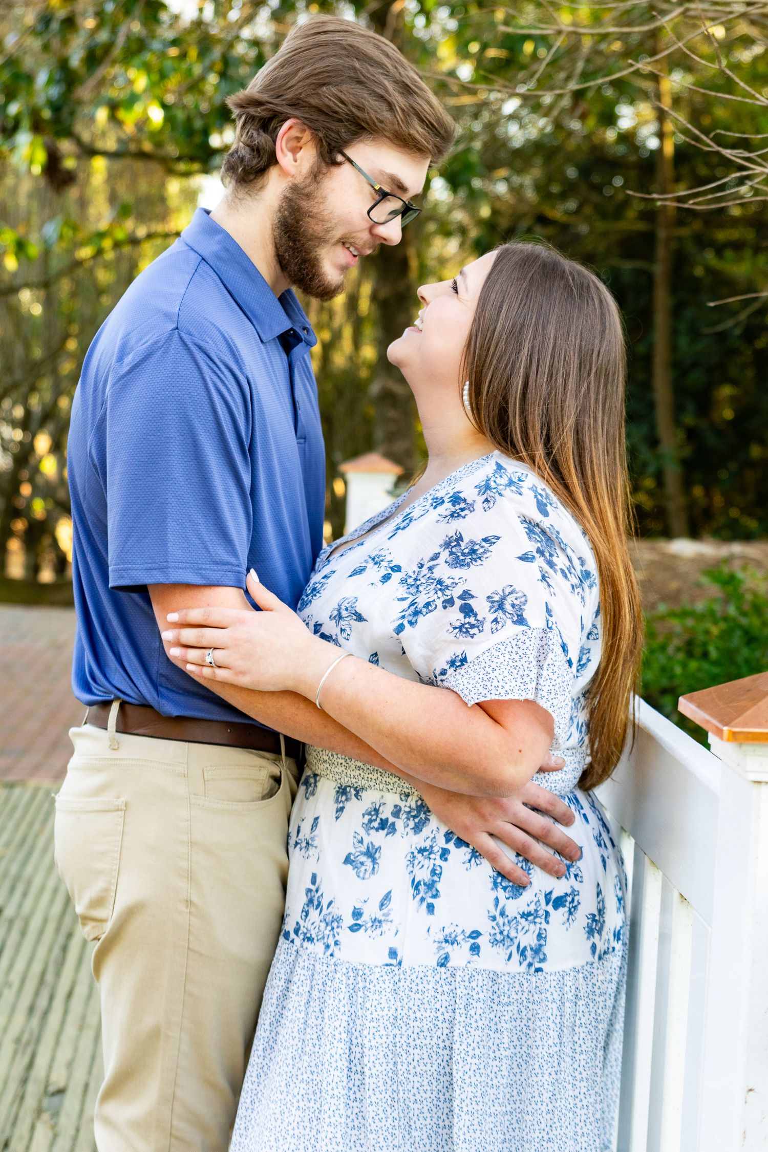A couple in blue and white outfits shares a romantic moment embracing on an outdoor deck surrounded by greenery.