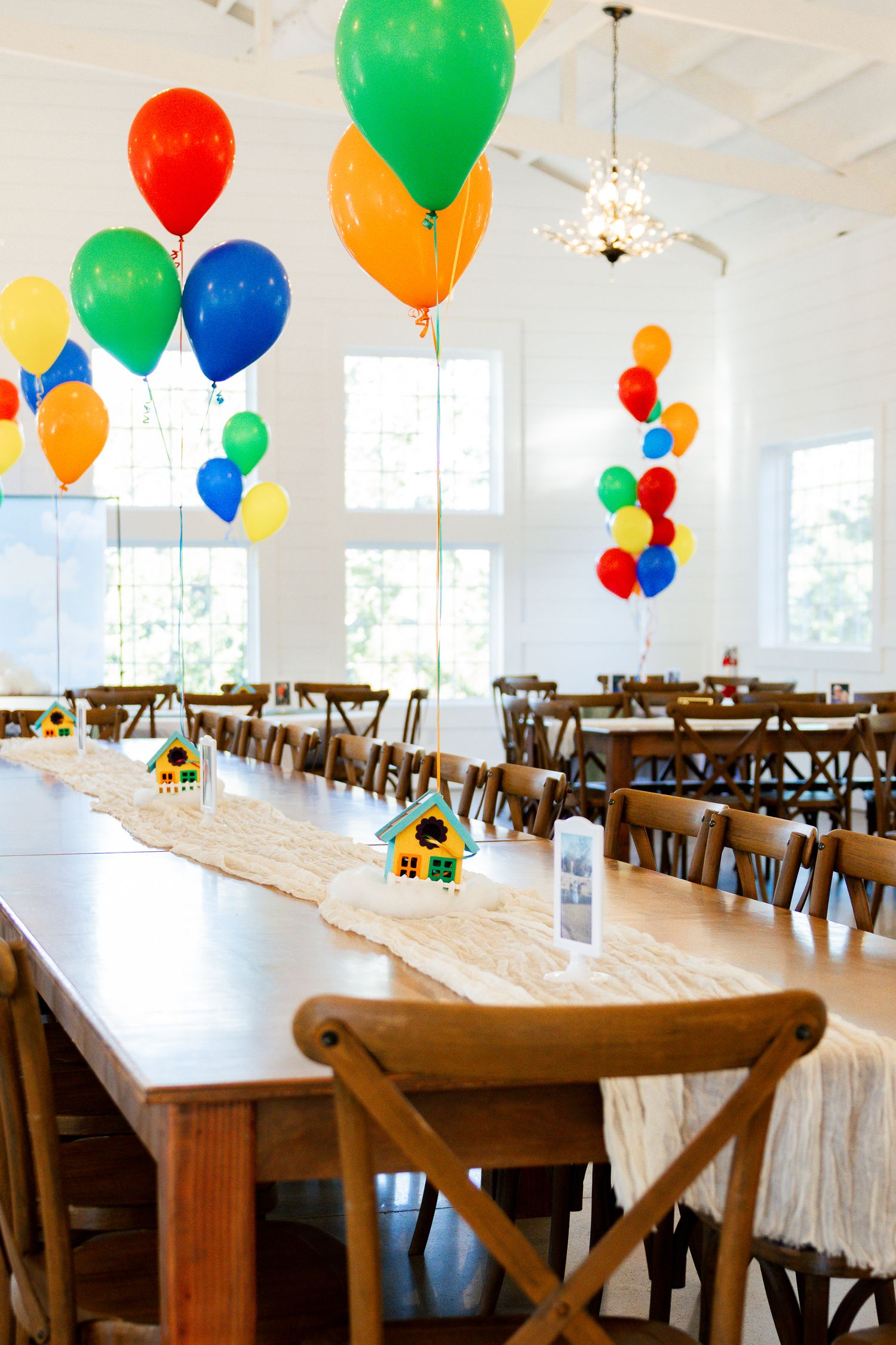 Colorful party balloons float above wooden dining tables in a bright white event space decorated for a celebration.