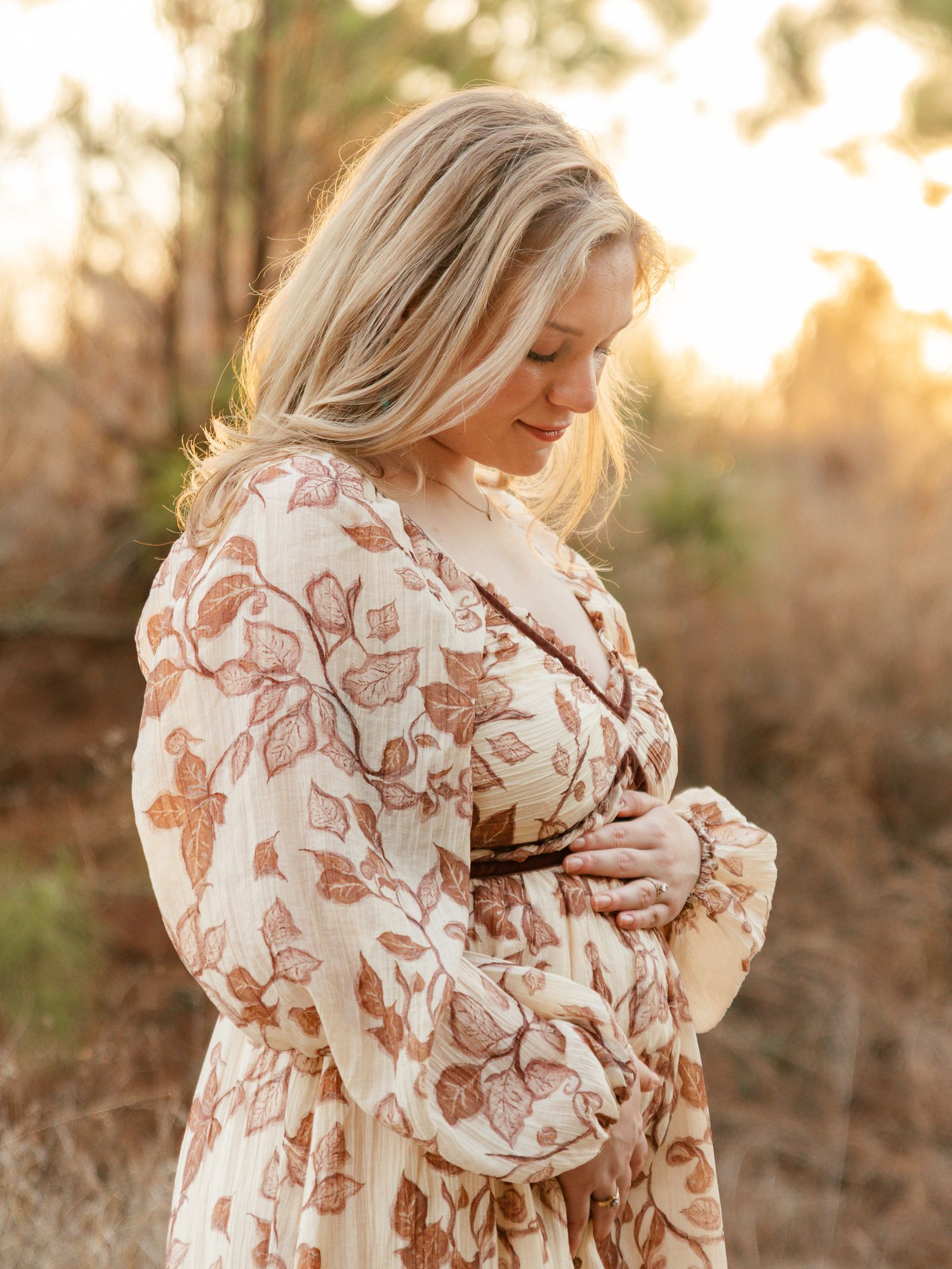 A pregnant woman in a floral dress poses outdoors during golden hour sunset with her hands on her baby bump.