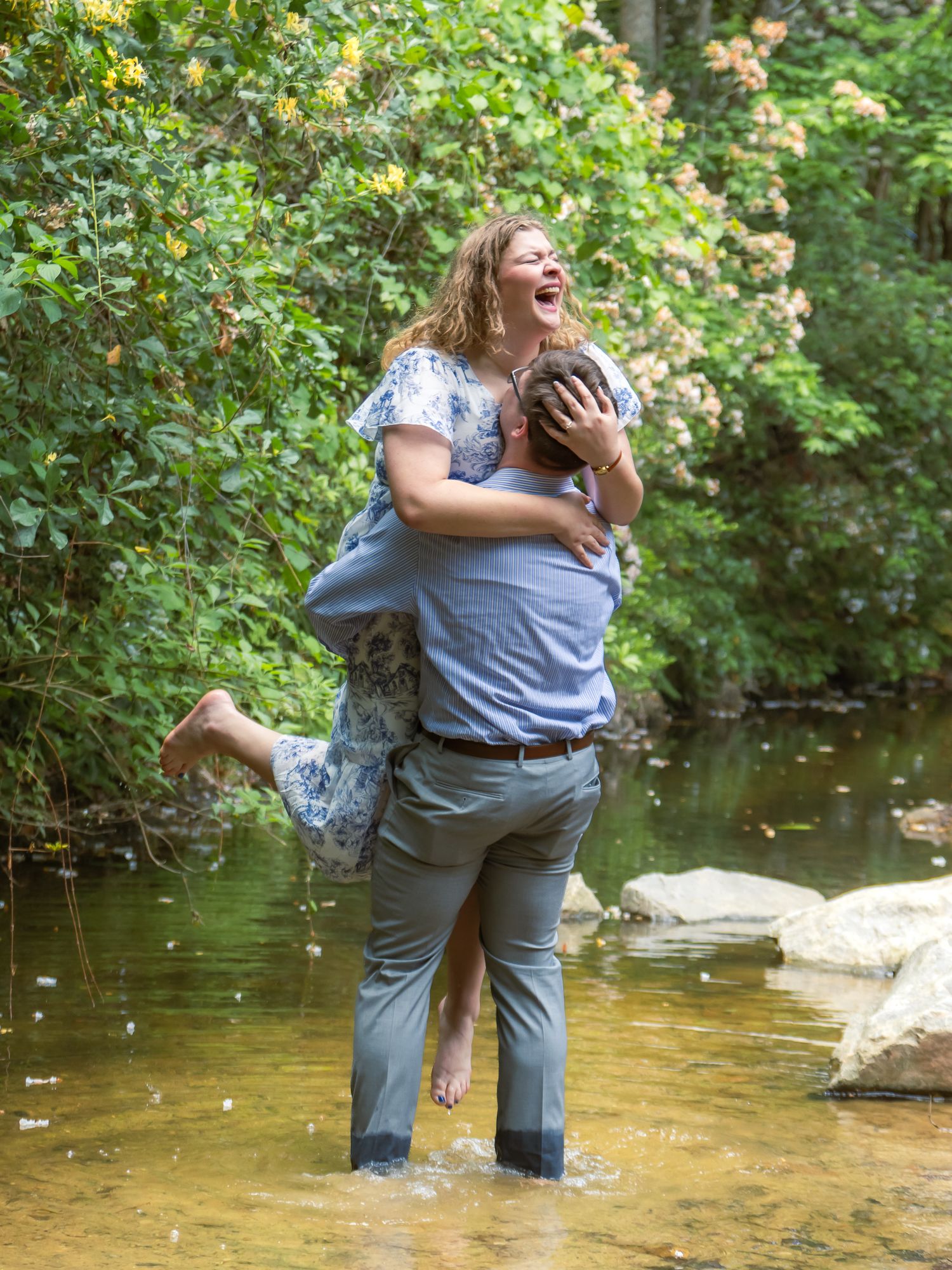 A joyful couple laughs together as a man lifts a woman in a shallow creek surrounded by lush green trees.