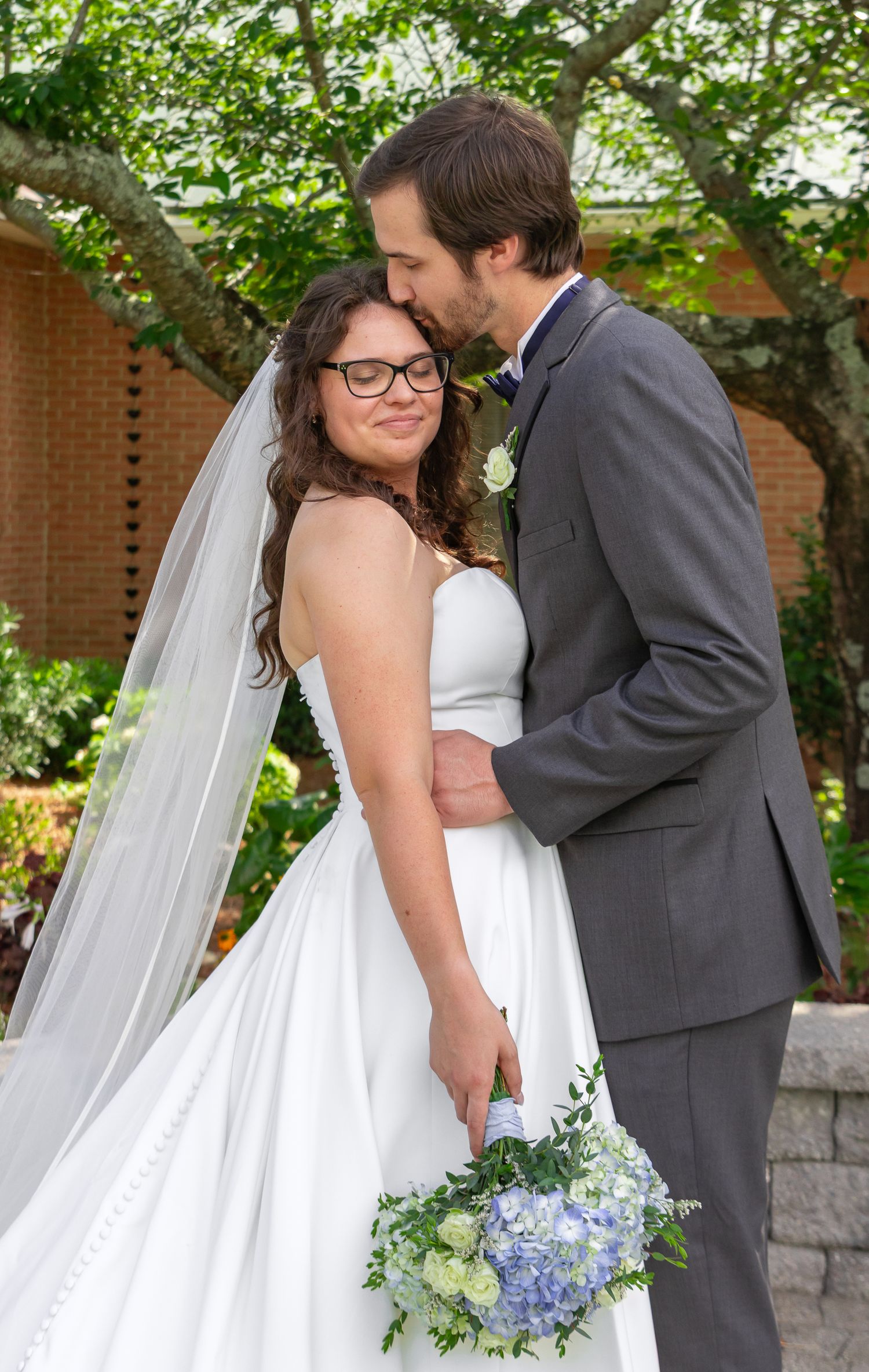 Bride in white gown with veil holds blue bouquet as groom in gray suit kisses her forehead outdoors.