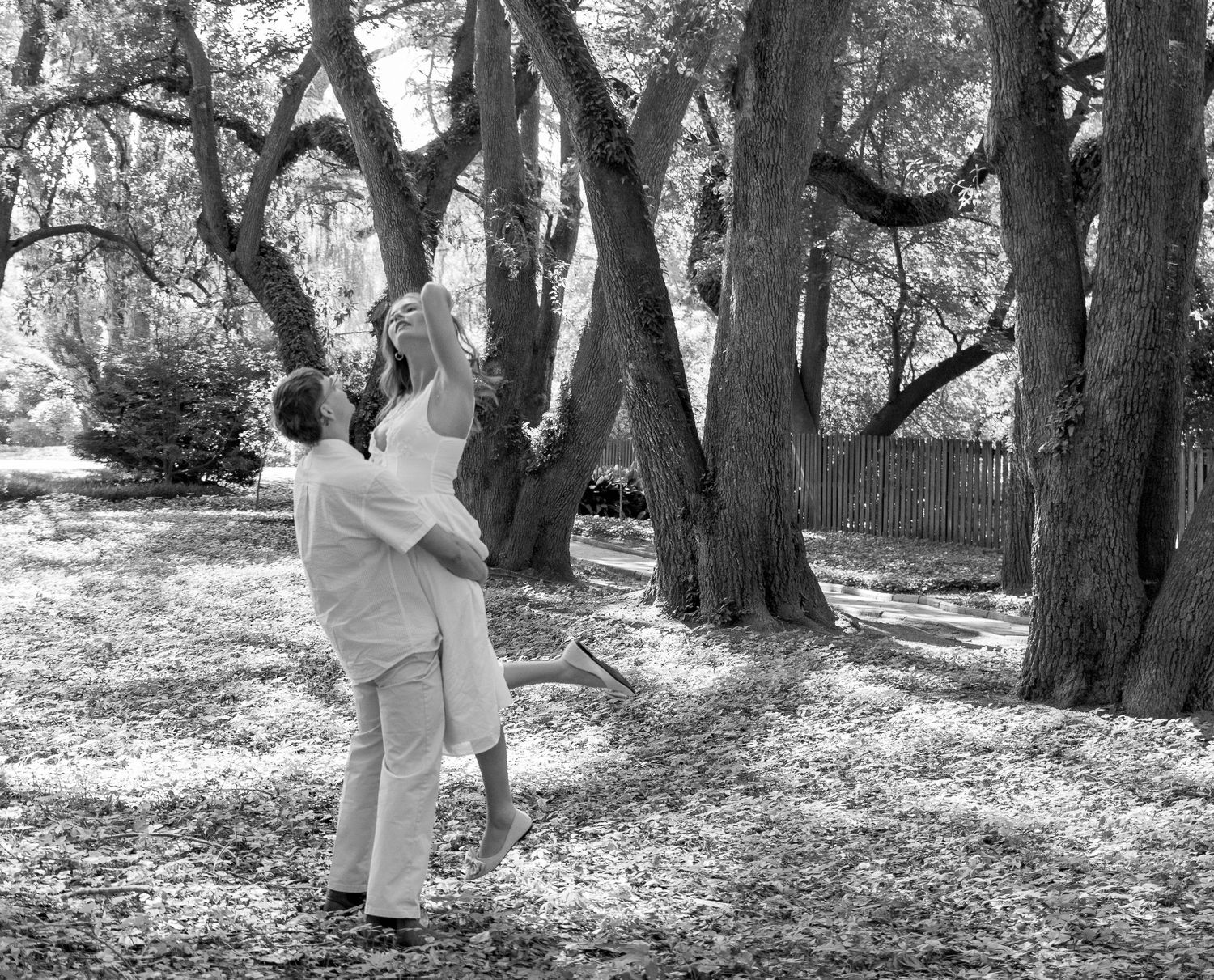 Black and white photograph of an engaged couple dancing beneath large trees in Aiken, South Carolina.