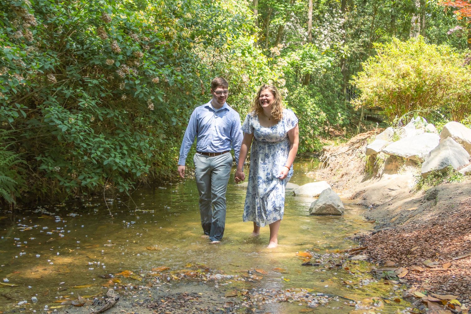 Couple walking hand in hand through a shallow creek surrounded by lush green trees and rocks.