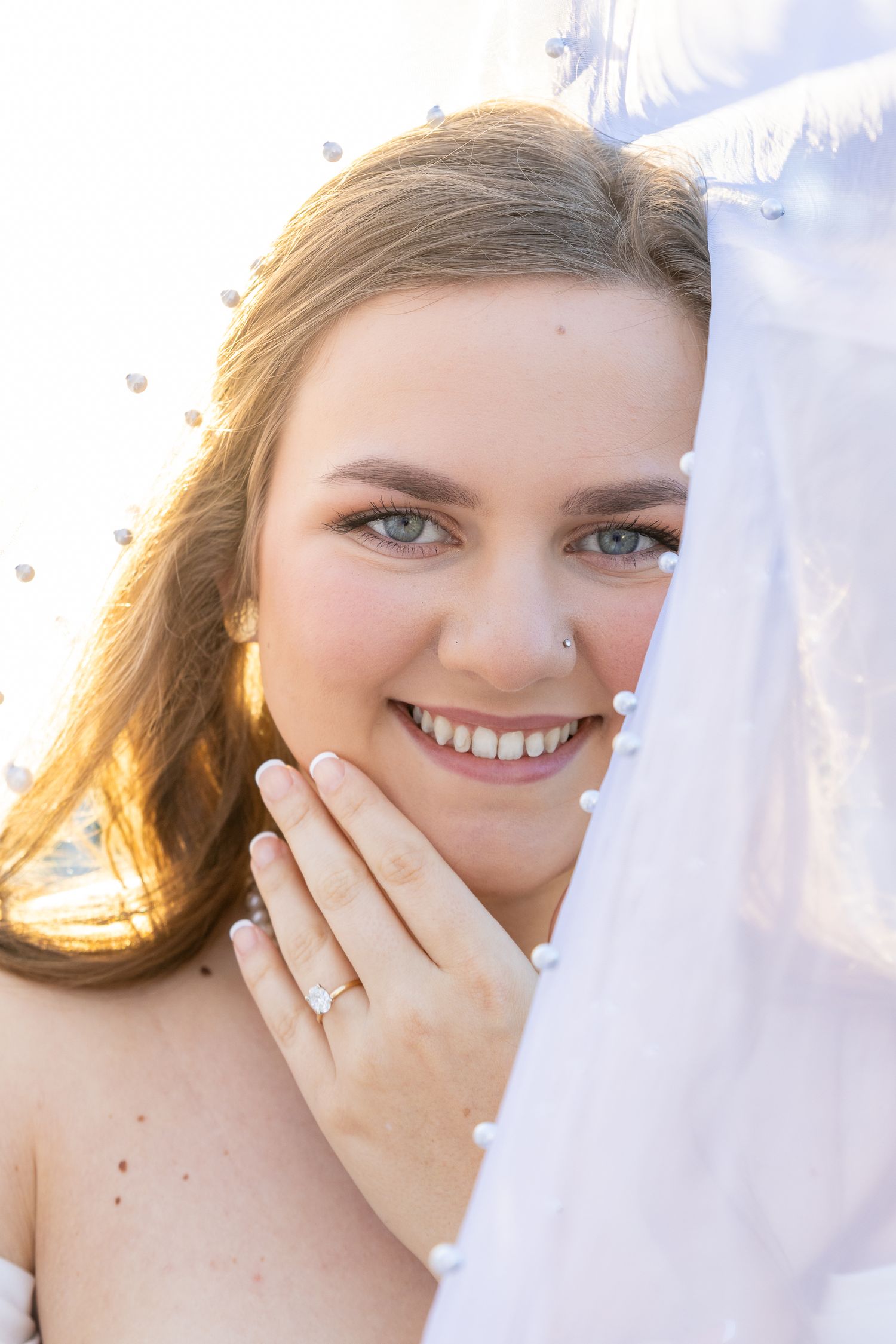 Smiling bride with blue eyes peeking behind pearl-trimmed veil displaying engagement ring in sunlight.