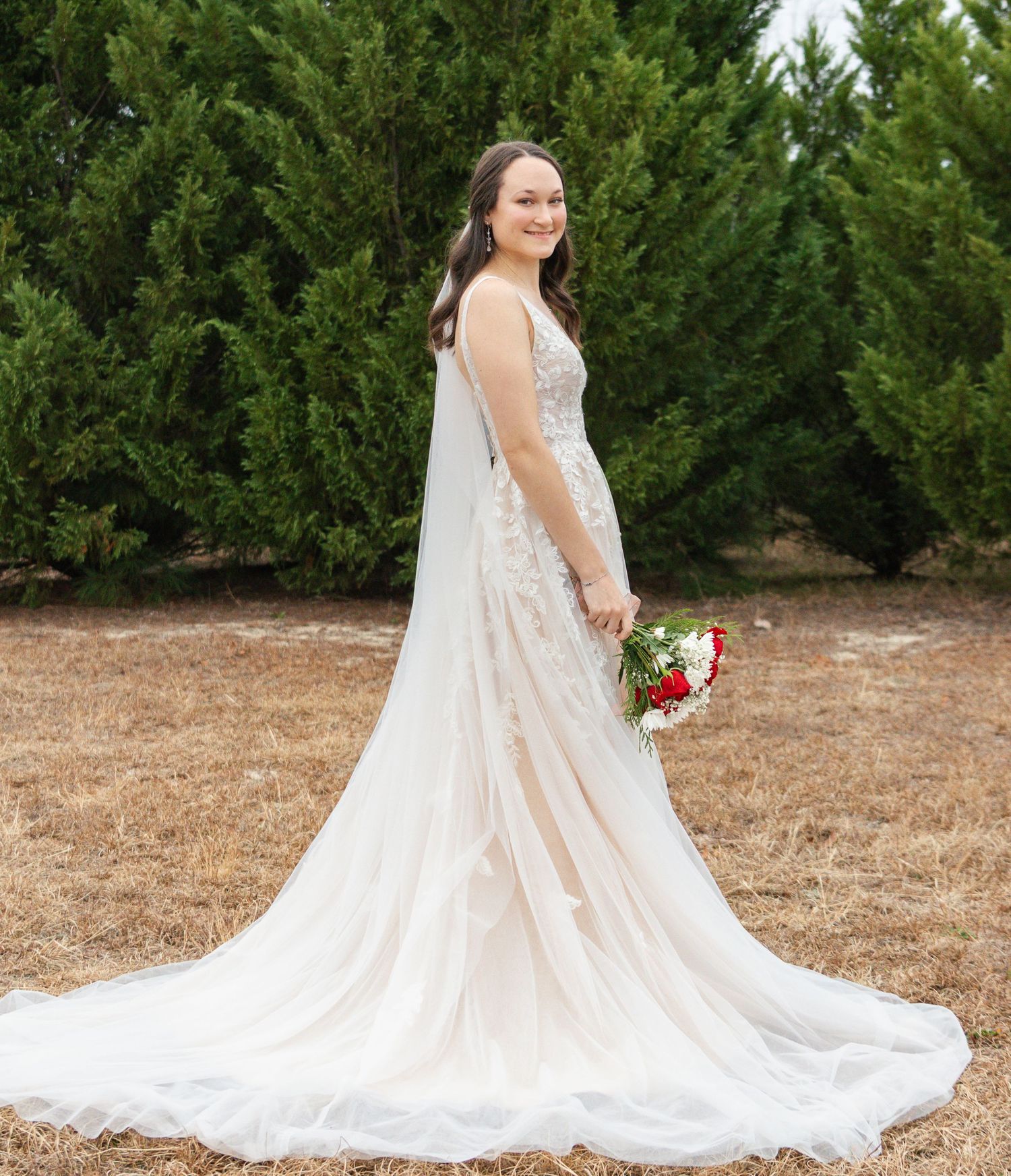 Bride in lace A-line wedding gown with long veil holding red and white bouquet outdoors.